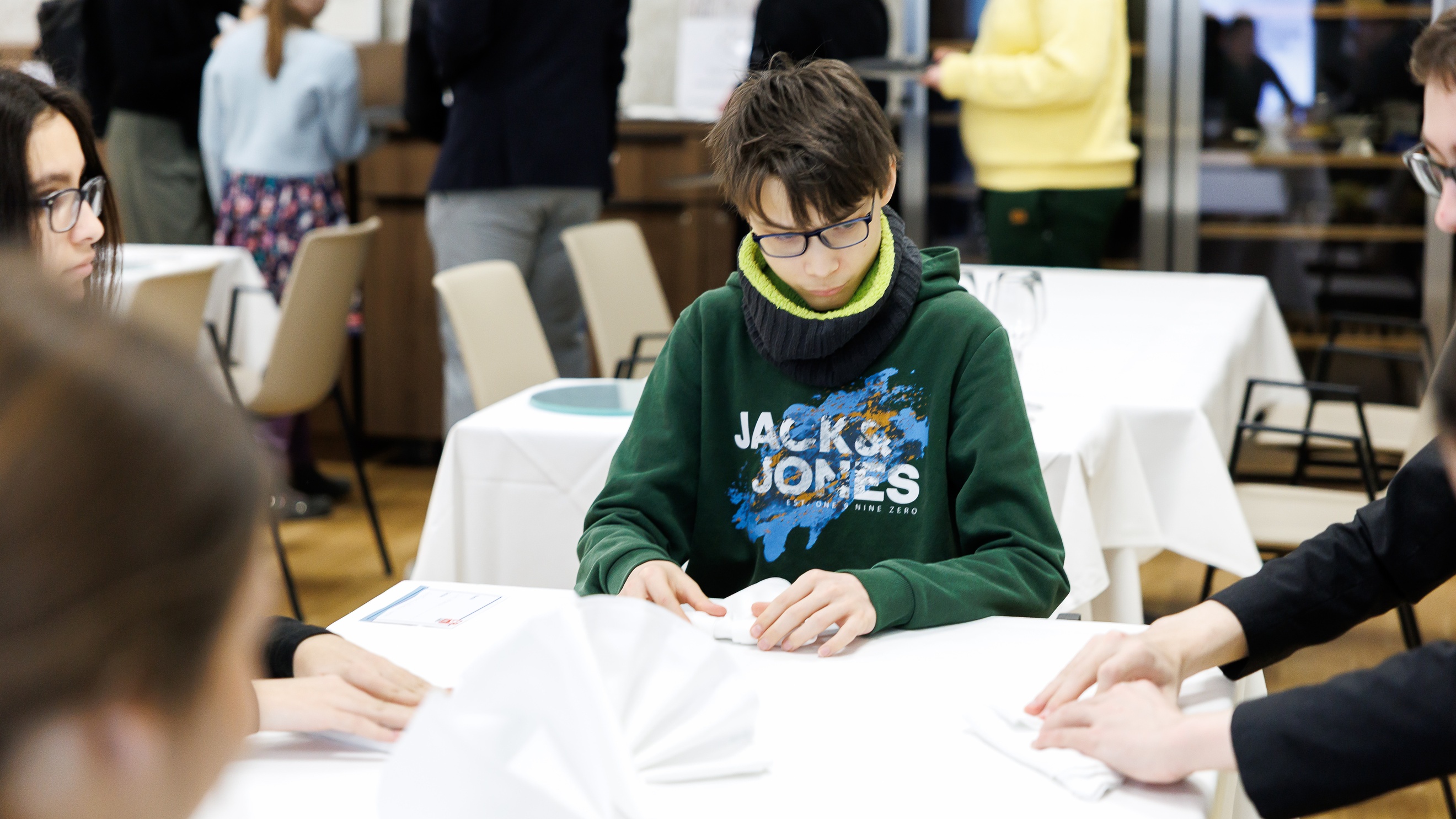 Um einen Tisch mit einem weißen Tischtuch sitzen vier jugendliche Personen. Jede Person faltet eine weiße Serviette