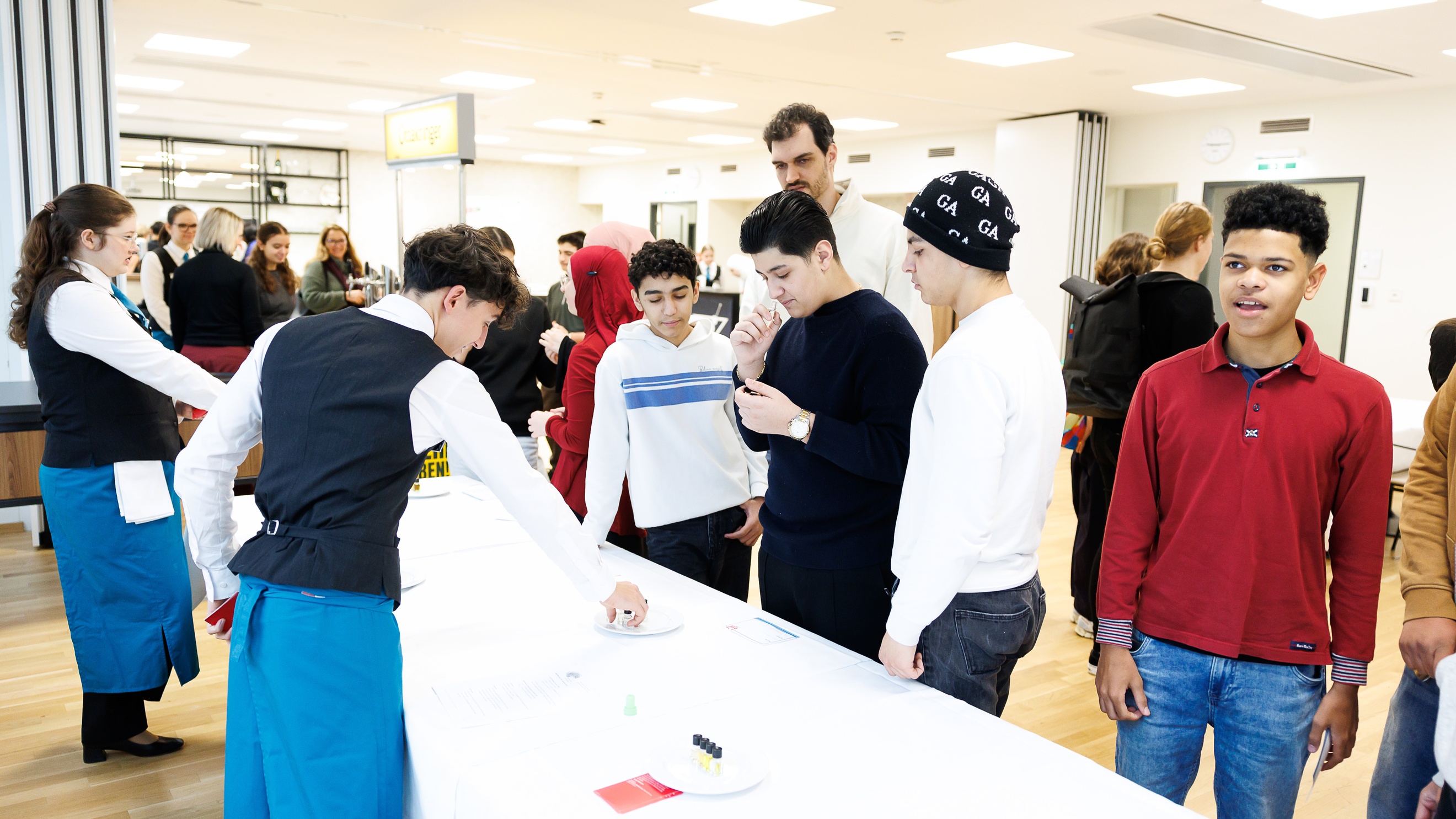 In einem  Raum stehen an einem langen Tisch auf der einen Seite zwei Personen in weißen Hemden, Gilets und Schürzen. Auf der anderen Seite des Tisches stehen jugendliche Personen