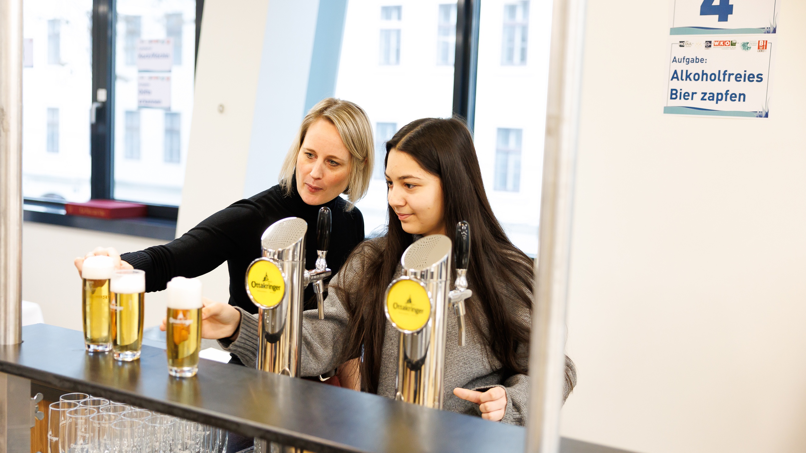 Eine Person mit geschlossenen, dunklen Haaren steht neben einem Zapfhahn. Neben ihr steht eine Person mit kinnlangen, blonden Haaren. Jede Person stellt jeweils Biergläser auf dem Tresen ab