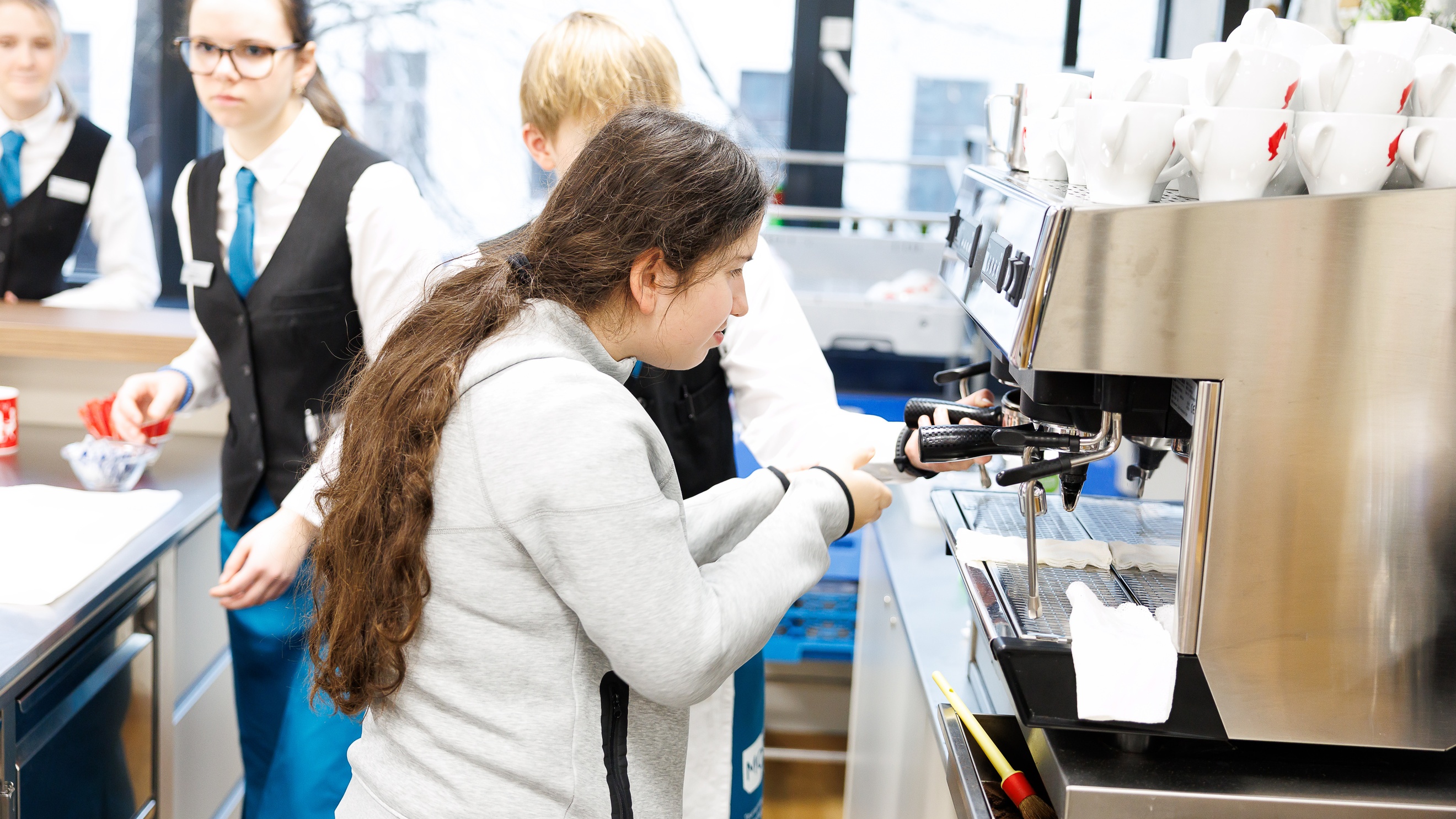 Seitliche Aufnahme einer jugendlichen Person, die eine Espressomaschine bedient. Neben ihr steht eine weitere Person, die ihr hilft
