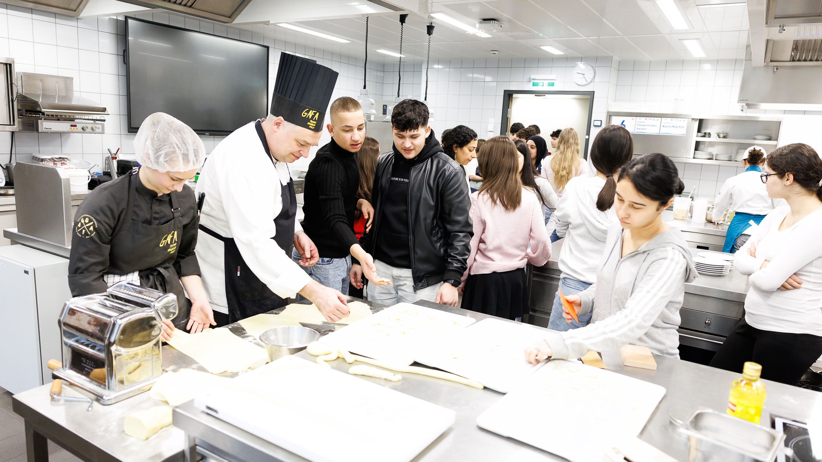 In einer Gastronomieküche stehen an verschiedenen Stationen mehrere Personen. Im Vordergrund stehen drei jugendliche Personen sowie zwei Personen in Kochkleidung. Auf dem Metalltisch vor ihnen liegt Teig