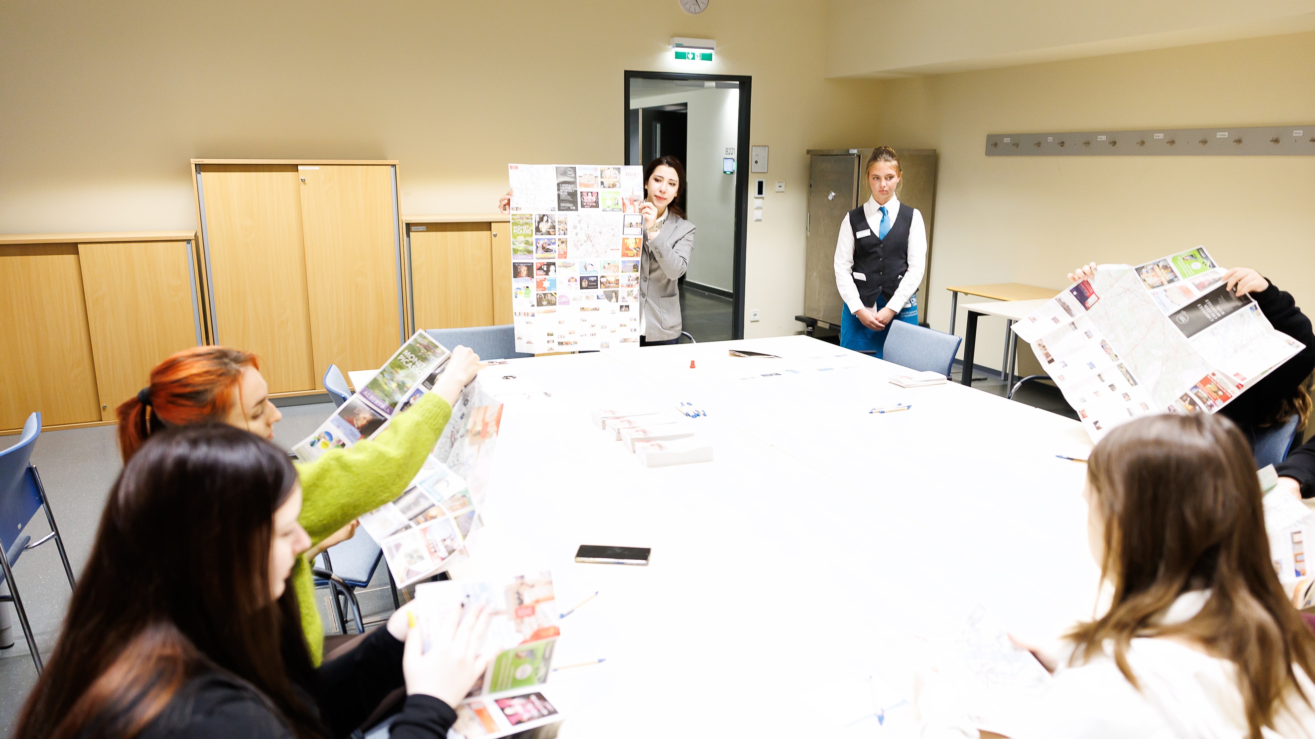 Um einen großen Tisch sitzen vier Personen. Zwei weitere Personen stehen daneben. Die sitzenden Personen sowie eine der stehenden Personen halten jeweils eine Broschüre