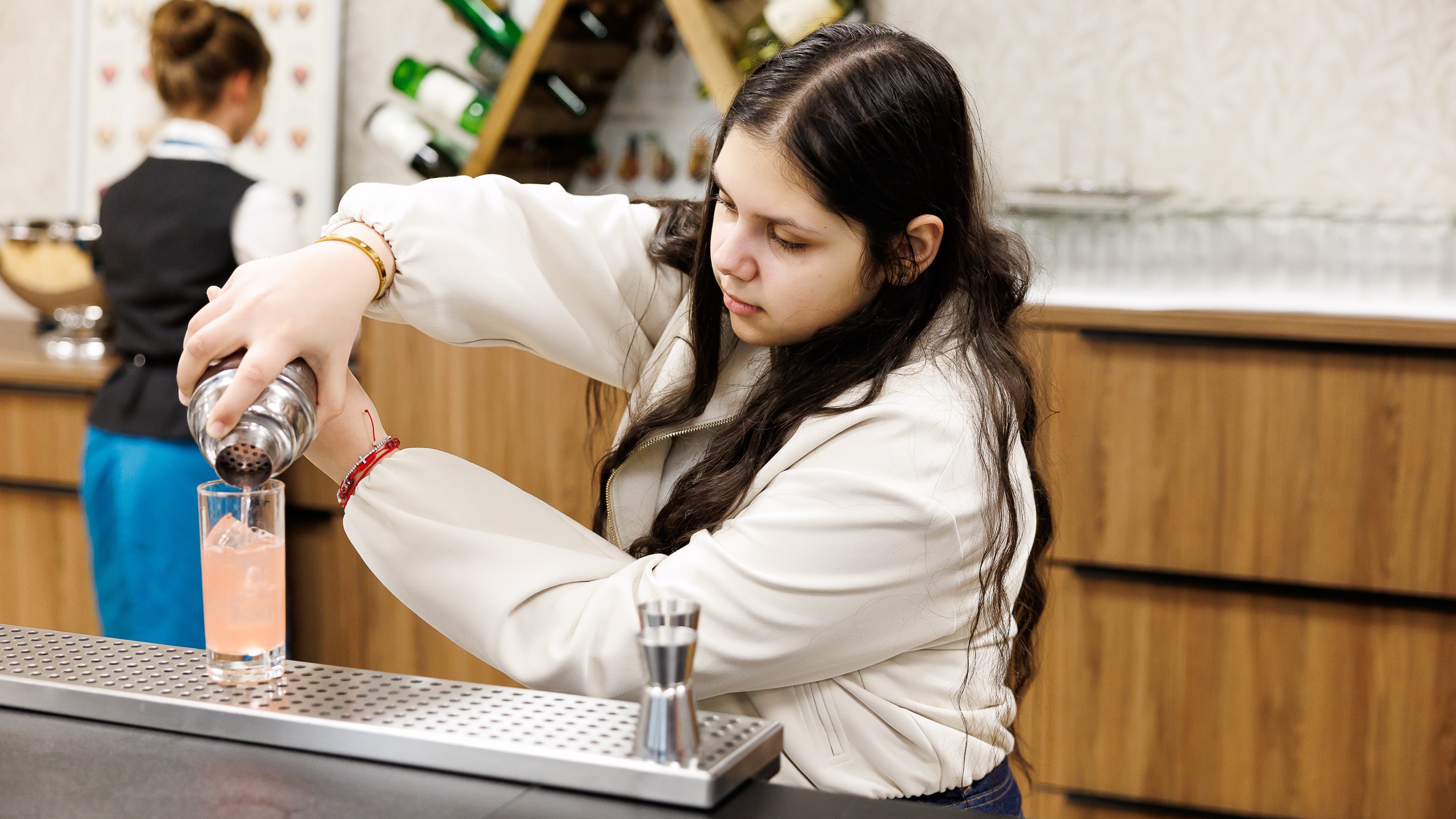 Eine jugendliche Person steht hinter einem Tresen und gießt aus einem Cocktailshaker ein hellrosa Getränk in ein Glas