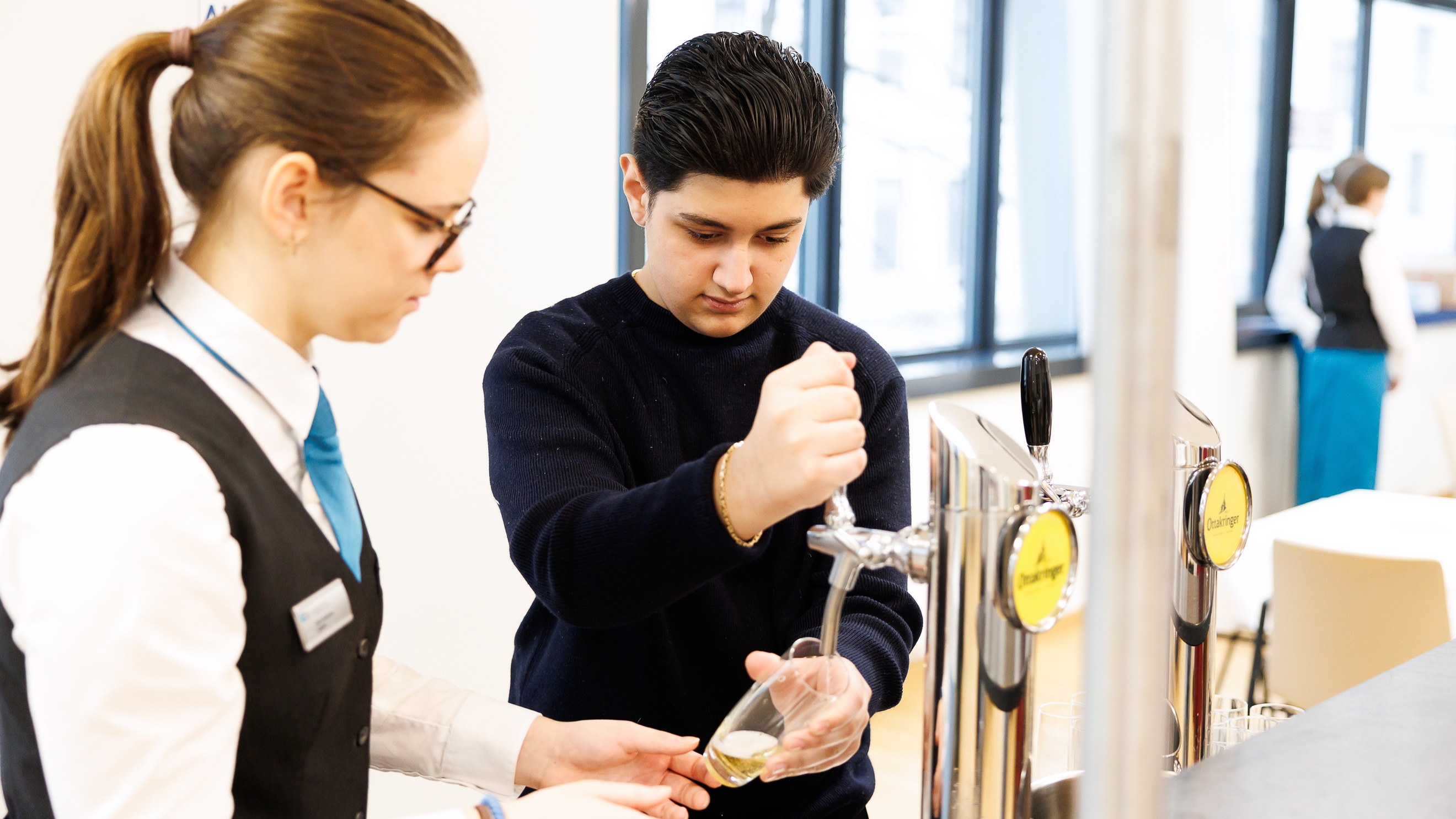 Eine Person mit dunklen, kurzen Haaren zapft aus einem Zapfhahn Bier. Neben ihr steht eine Person mit geschlossenen Haaren, Brille, weißer Bluse, Gilet und Krawatte, die der anderen Person hilft
