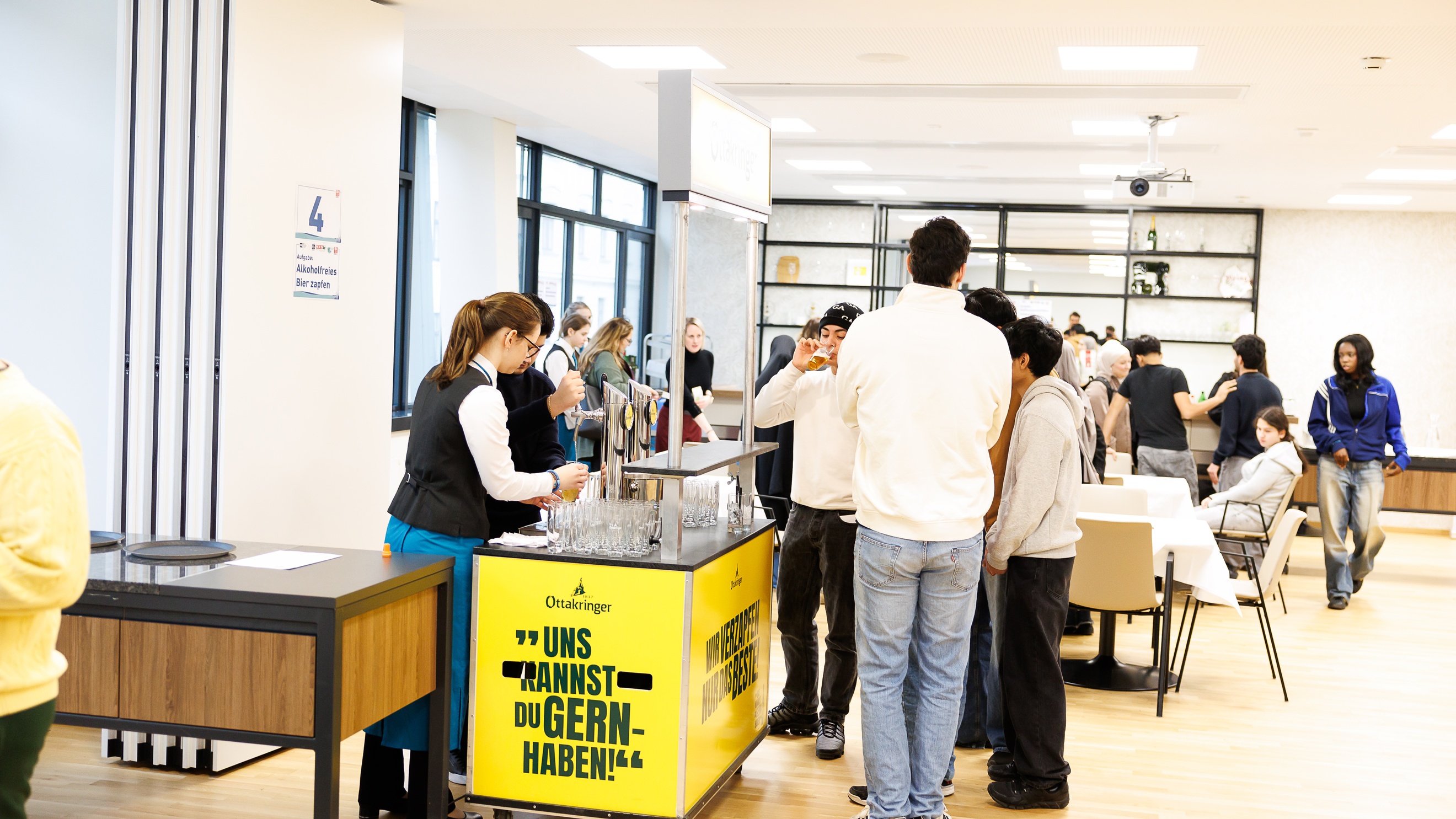 In einem Raum stehen hinter einem mobilen Zapfhahn zwei Personen. Daneben stehen Personen. Im Hintergrund sind mehrere Tische, an denen Personen sitzen