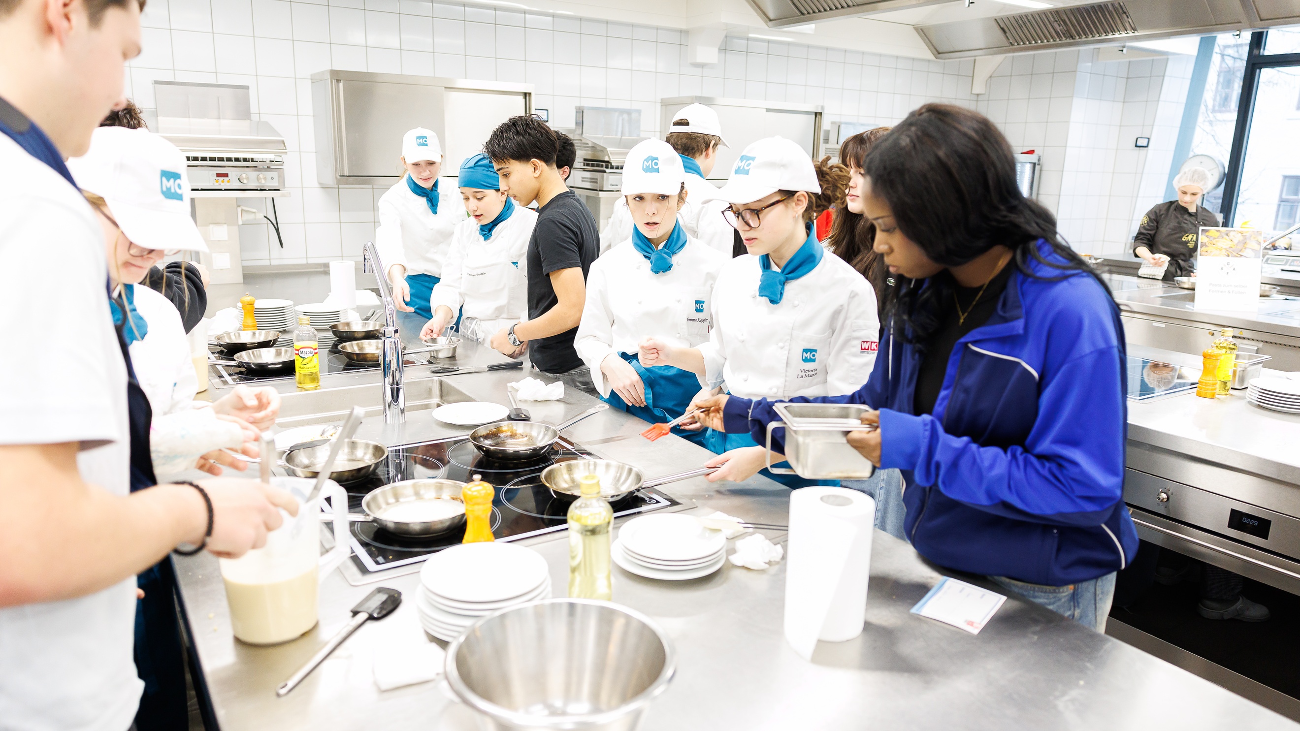 In einer Gastronomieküche stehen an verschiedenen Stationen mehrere Personen. Im Vordergrund stehen an einem Ceranfeld vier Personen, die jeweils vor sich eine Pfanne stehen haben. Neben zwei der Personen steht eine weitere Person in weißer Kochkleidung