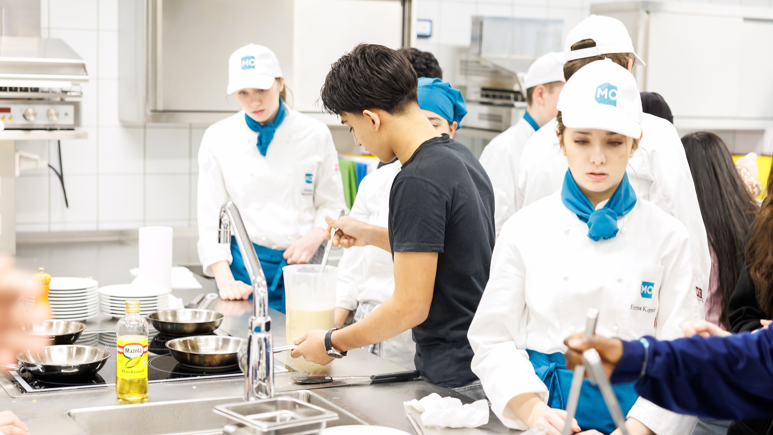 In einer Gastronomieküche stehen an verschiedenen Stationen mehrere Personen. Unter anderem stehen an einem Ceranfeld eine jugendliche Person, die Teig anrührt sowie eine Person in weißer Kochkleidung