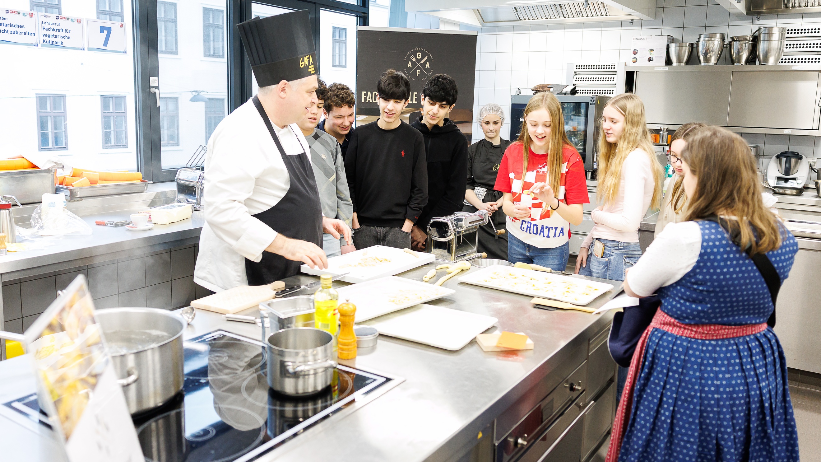 In einer Gastronomieküche stehen an verschiedenen Stationen mehrere Personen. Im Vordergrund stehen sieben jugendliche Personen sowie eine in Kochkleidung. Auf dem Metalltisch vor ihnen liegt Teig