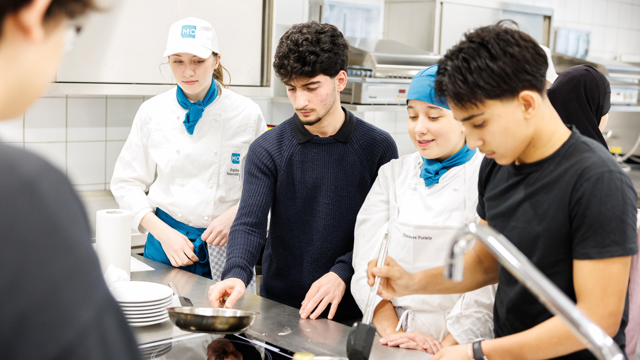 In einer Gastronomieküche stehen an einem Ceranfeld zwei jugendliche Personen sowie zwei Personen in Kochkleidung. Die jugendlichen Personen haben jeweils vor sich eine Pfanne