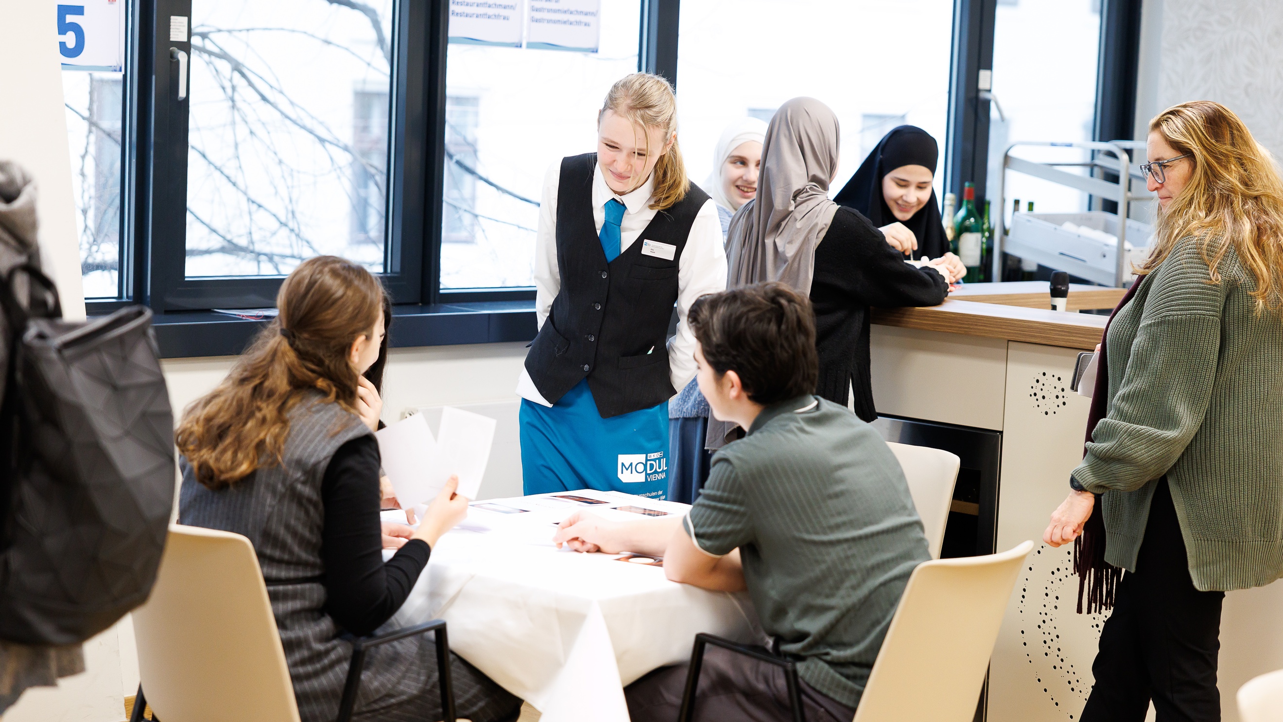 Um einen Tisch sitzen drei Personen. Neben ihr steht eine Person in weißer Bluse, mit Gilet, Krawatte und blauer Schürze