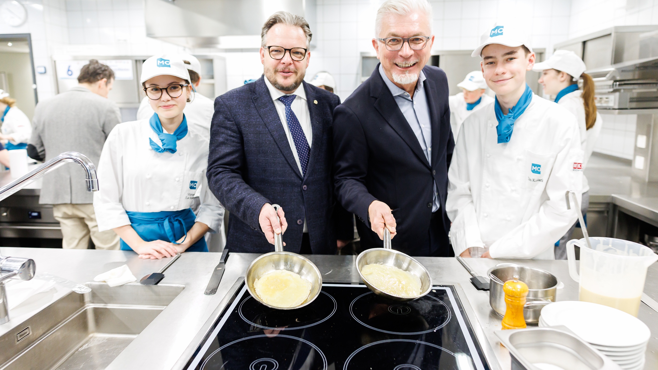 Porträt von zwei Personen in dunklen Sakkos und mit Bart, die hinter einem Ceranfeld stehen und jeweils eine Pfanne halten. Neben ihnen stehen zwei jugendliche Personen in Kochkleidung. Im Hintergrund sind weitere Personen
