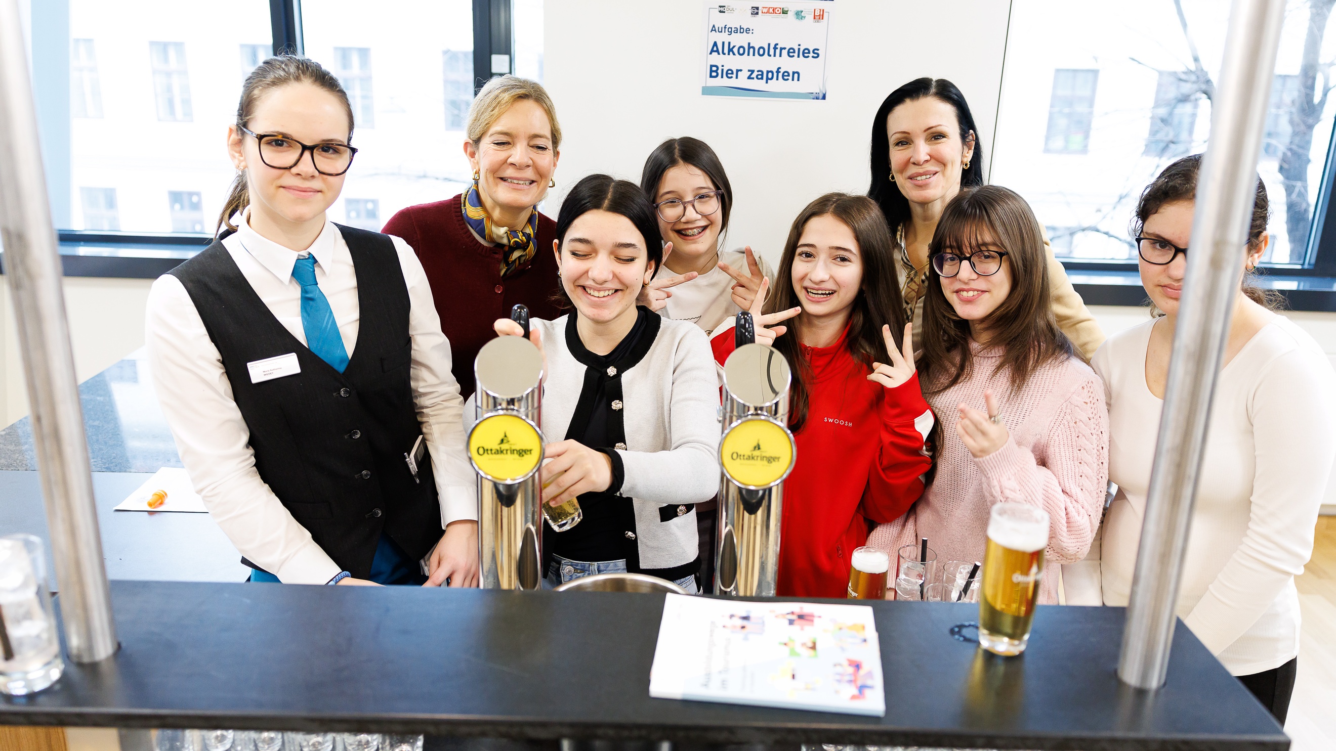 Porträt von fünf jugendlichen Personen, zwei erwachsenen Personen und einer Person in hellem Hemd, Gilet und Krawatte, die nebeneinander hinter einem Tresen stehen. Eine der jugendlichen Personen bedient einen Zapfhahn
