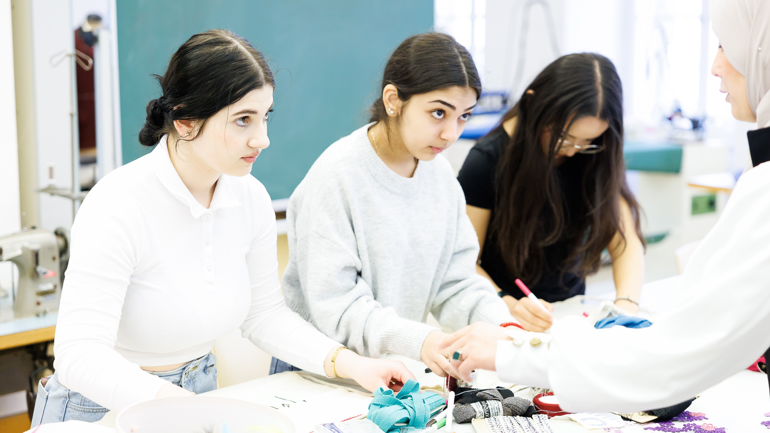 Drei Teenager stehen nebeneinander an einem Tisch. Vor ihnen sind verschiedene Stoffe. Ein Teenager malt mit einem roten Stift. Ihnen gegenüber steht eine erwachsene Person, die etwas erklärt