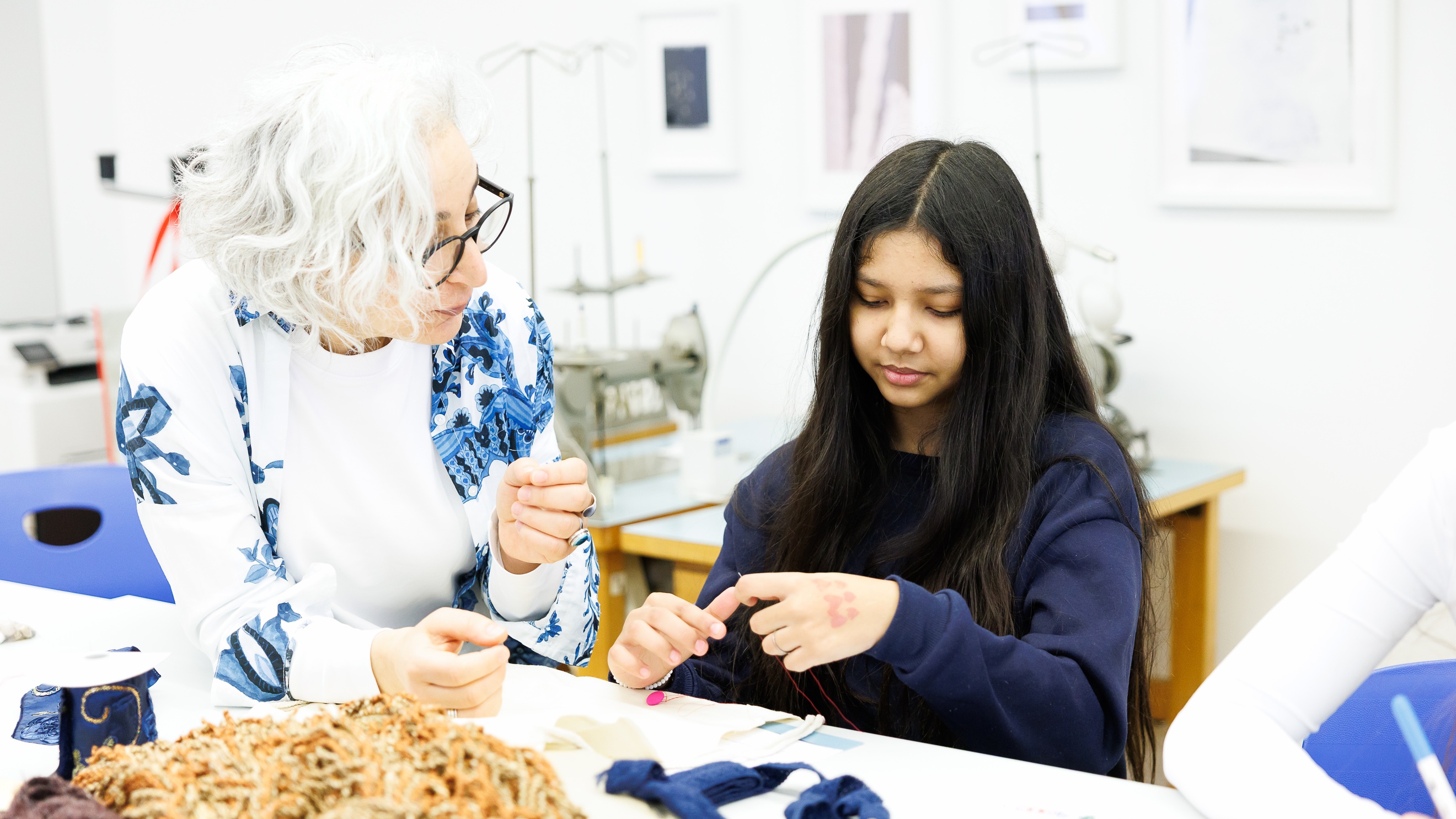 Ein Teenager sitzt an einem Tisch. Auf dem Tisch sind verschiedene Stoffe und Wollknäuel. Direkt neben dem Teenager ist eine Person mit weißen Haaren und Brille, die sich nach vorne beugt und etwas erklärt