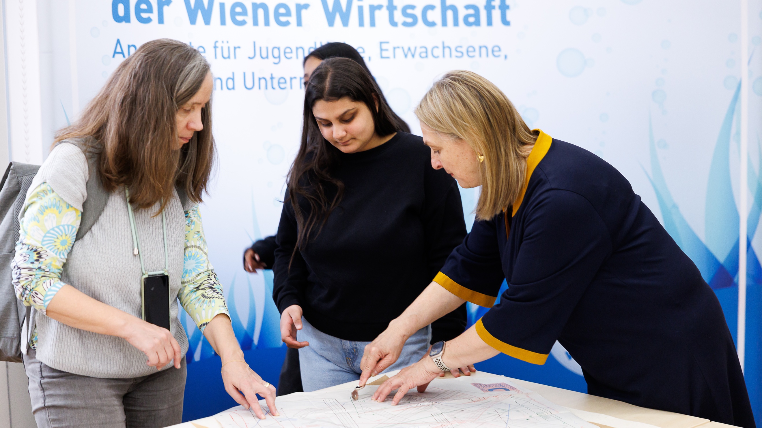 An einem Tisch stehen zwei erwachsene Personen und ein Teenager. Eine der erwachsenen Personen schneidet mit einem Messer ein Muster auf einem Blatt Papier aus. Die anderen beiden sehen zu