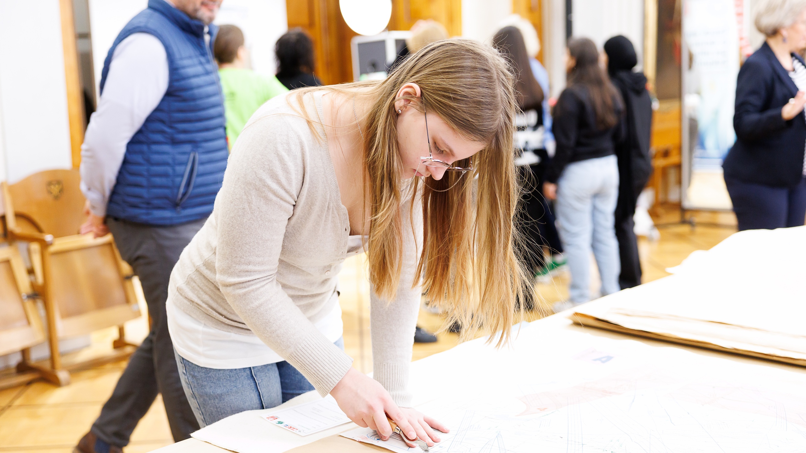 Ein Teenager mit Brille und offenen, hellen Haaren steht an einem Tisch und schneidet mit einem kleinen Messer ein Muster aus einem Blatt Papier aus. Im Hintergrund stehen weitere Teenager sowie erwachsene Personen