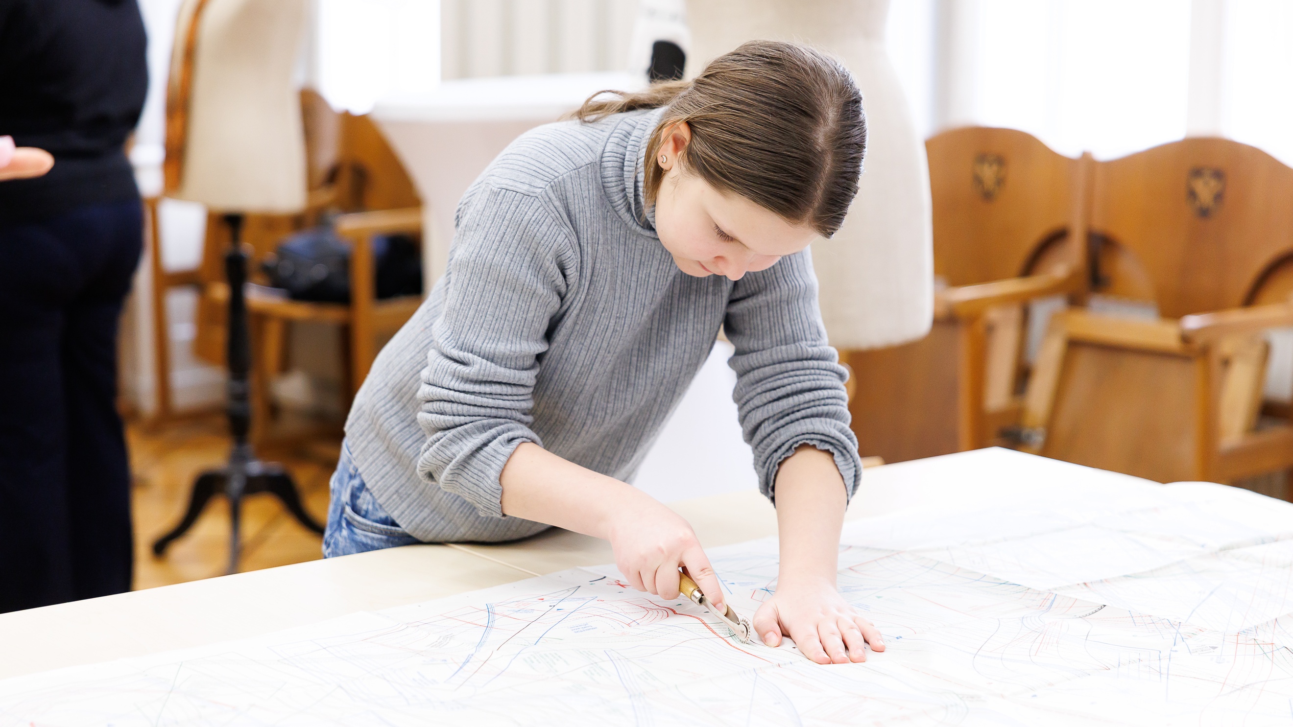Ein Teenager mit dunklen Haaren steht an einem Tisch und schneidet mit einem kleinen Messer ein Muster aus einem Blatt Papier aus. Im Hintergrund sind eine erwachsene Person sowie zwei versetzt stehende Modepuppen