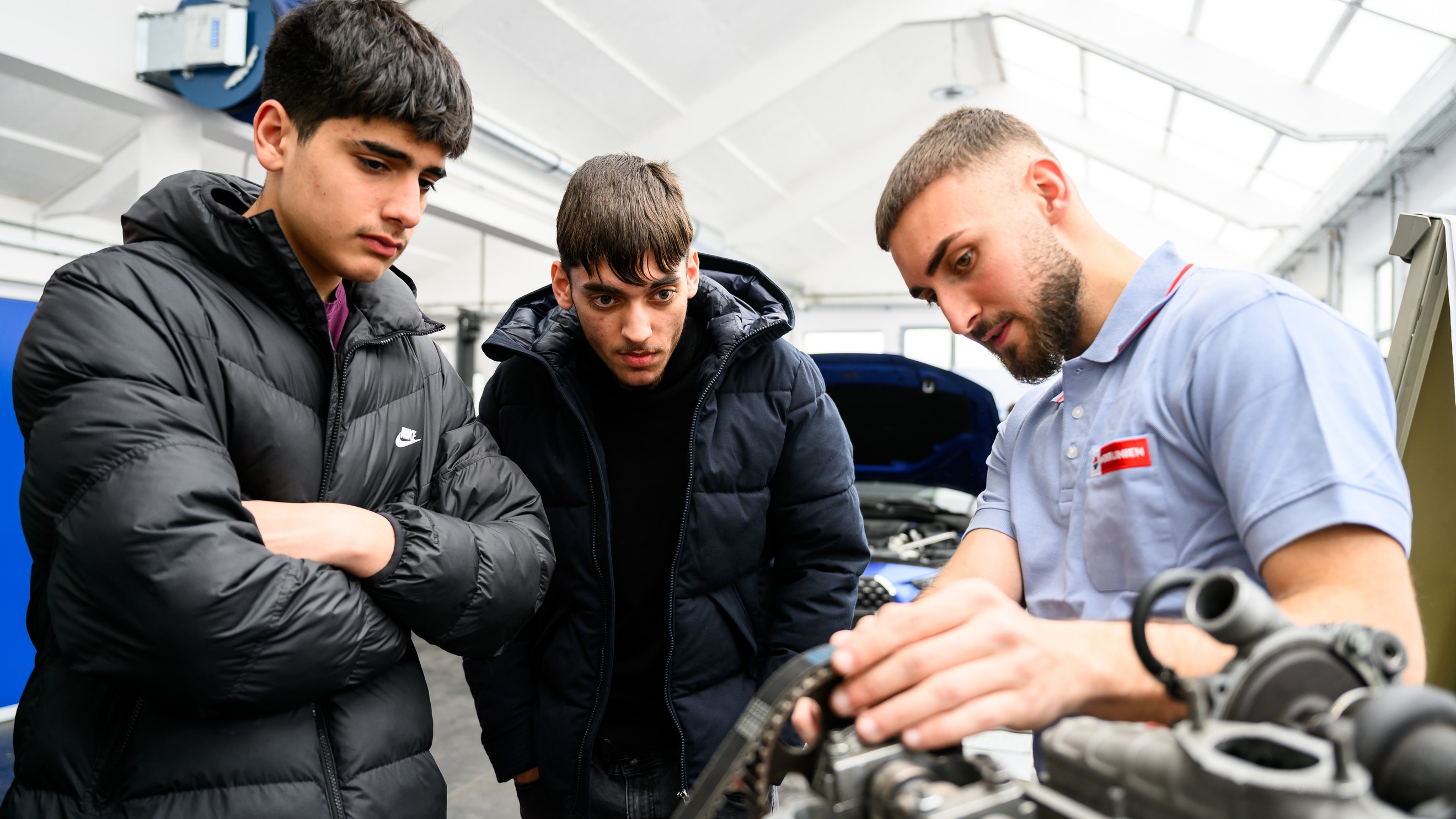 In einer Werkstatt stehen neben einem Motor eine erwachsene Person und zwei Teenager, die alle auf den Motor blicken. Im Hintergrund steht ein Auto mit geöffneter Motorhaube