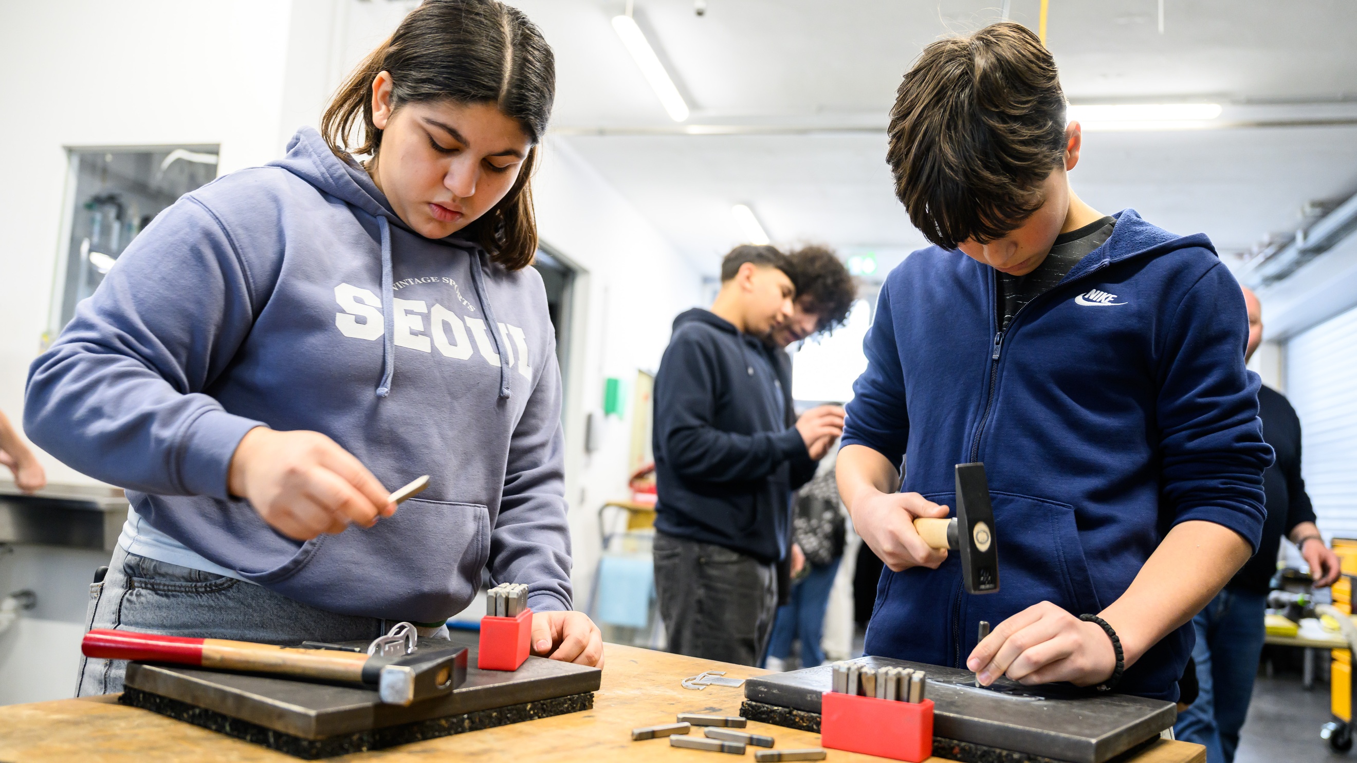 Ein Teenager steht an einem Tisch und schlägt mit einem Hammer einen breiten Nagel in eine MEtallplatte. Daneben steht ein weiterer Teenager, der einen Nagel hält