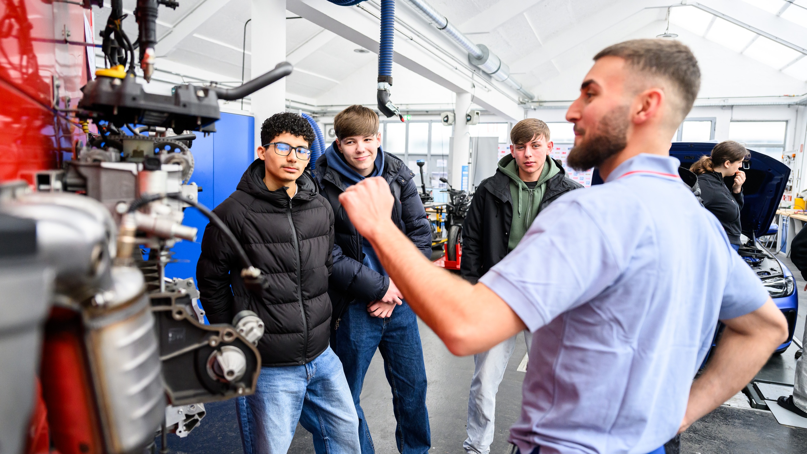 In einer Werkstatt steht eine Person neben einem Motor eines Fahrzeugs und redet. Der Person gegenüber stehen drei Teenager, die zuhören. Im Hintergrund steht ein Auto mit geöffneter Motorhaube