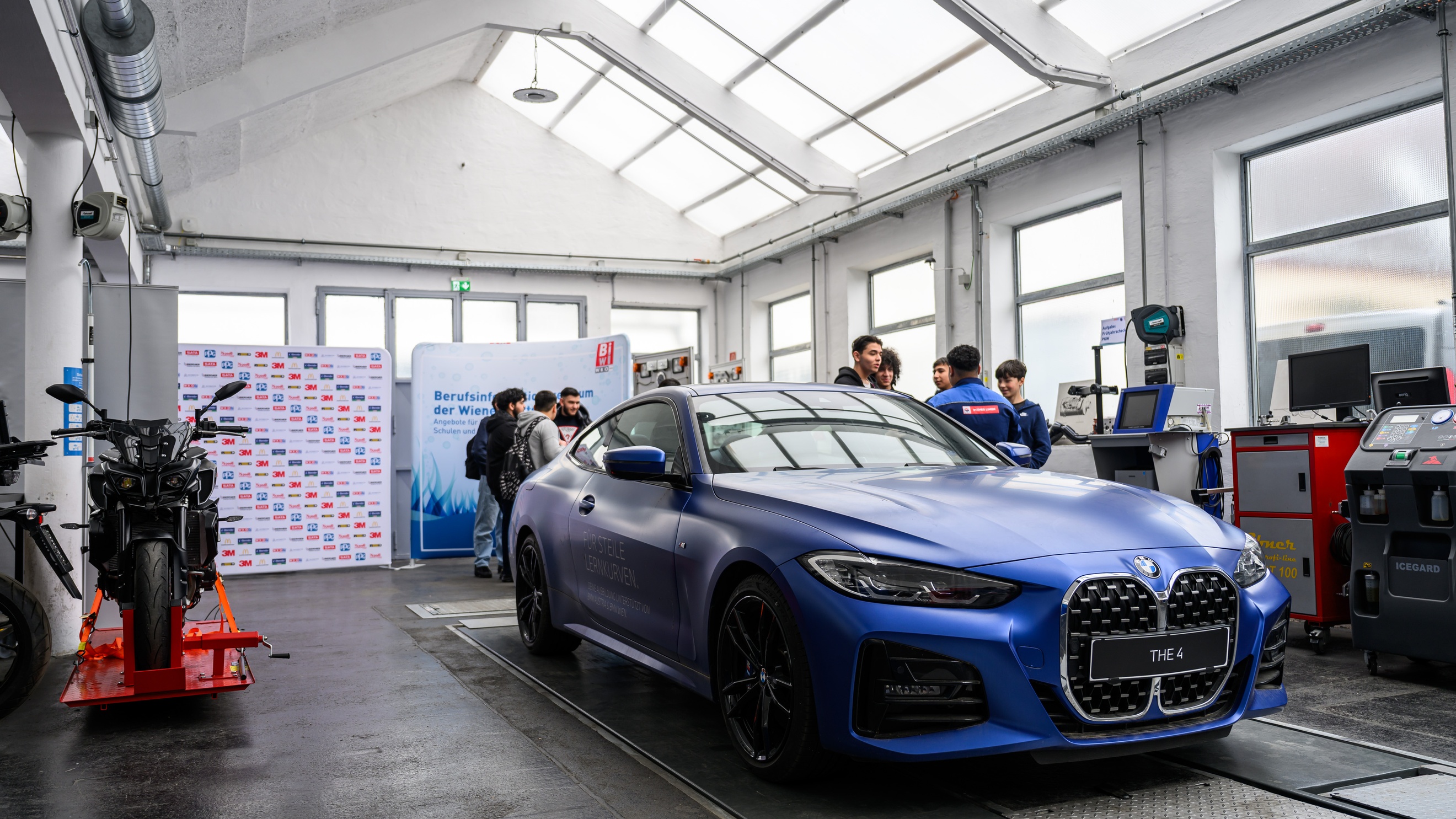 In einer Werkstatt steht ein blaues Auto. Daneben stehen mehrere Jugendliche