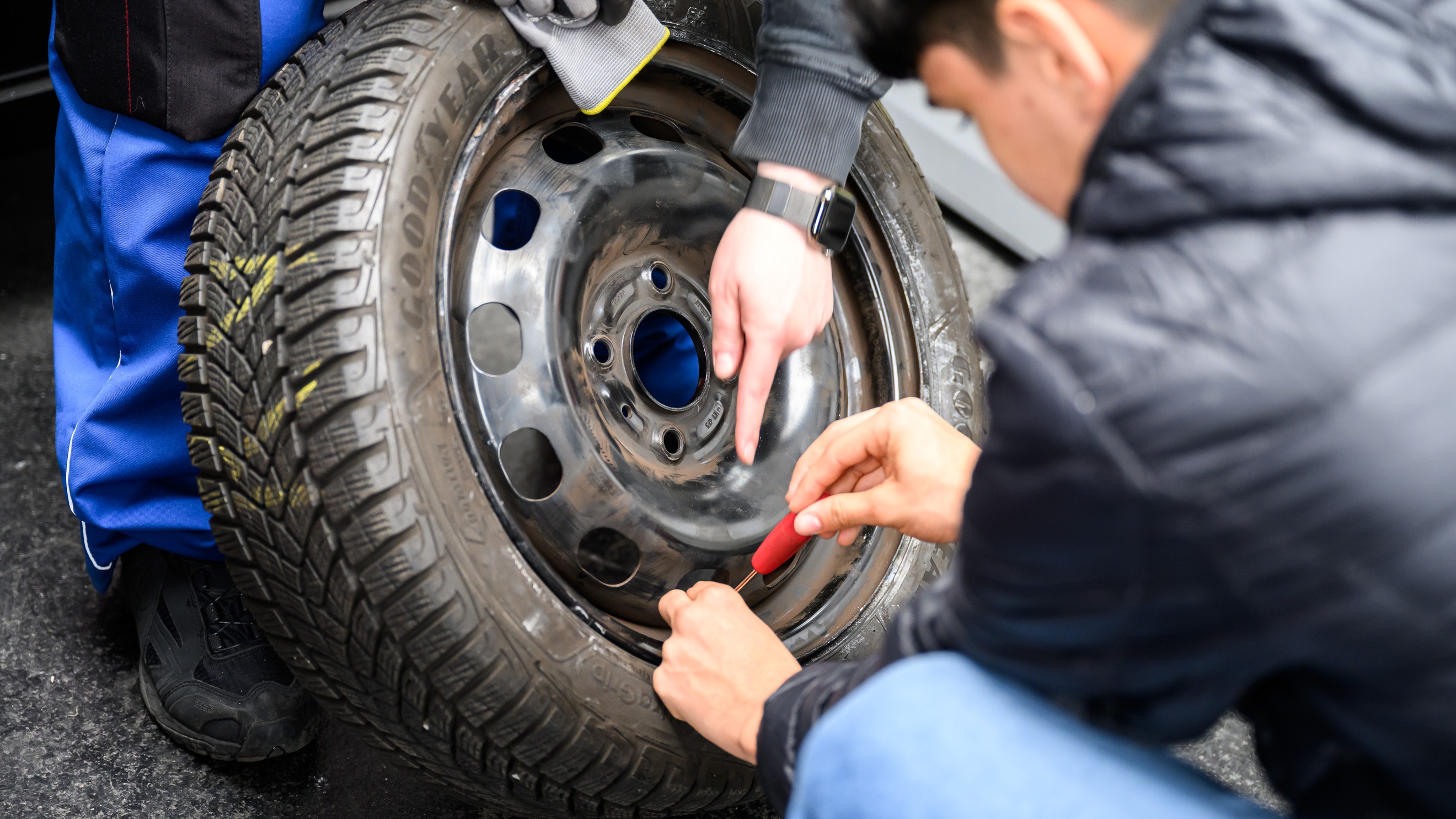 Nahaufnahme des Reifens eines Autos, bei dem eine Person eine Felge festschraubt. Die Hand einer anderen Person deutet auf die Felge