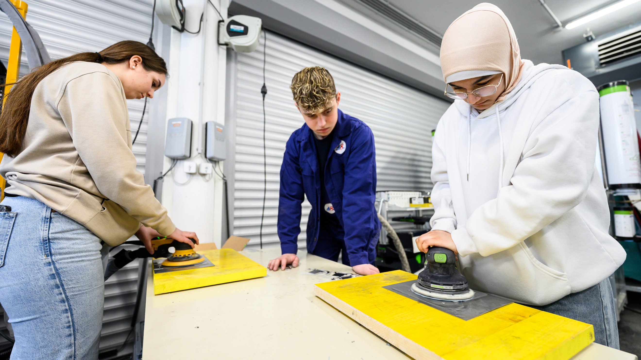 An einem Tisch in einer Werkstatt stehen zwei Personen, die auf Autoglasscheiben Saugknöpfe halten. Daneben steht eine Person in einem blauen Overall