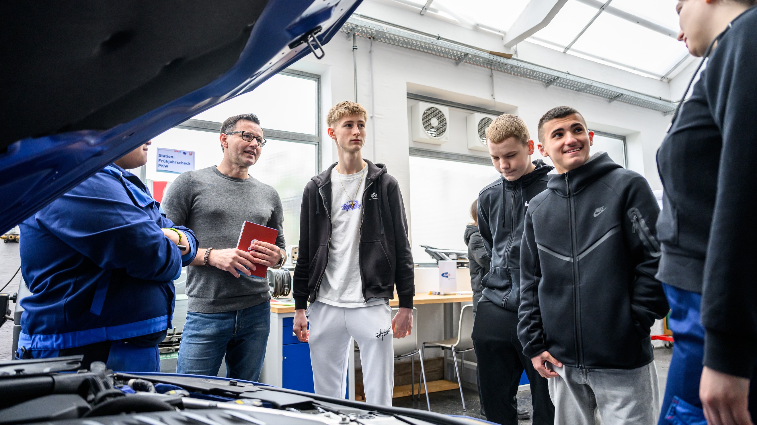 Neben einer geöffneten Motorhaube eines Fahrzeugs stehen drei erwachsene Personen und drei Teenager, die reden, in einer Werkstatt