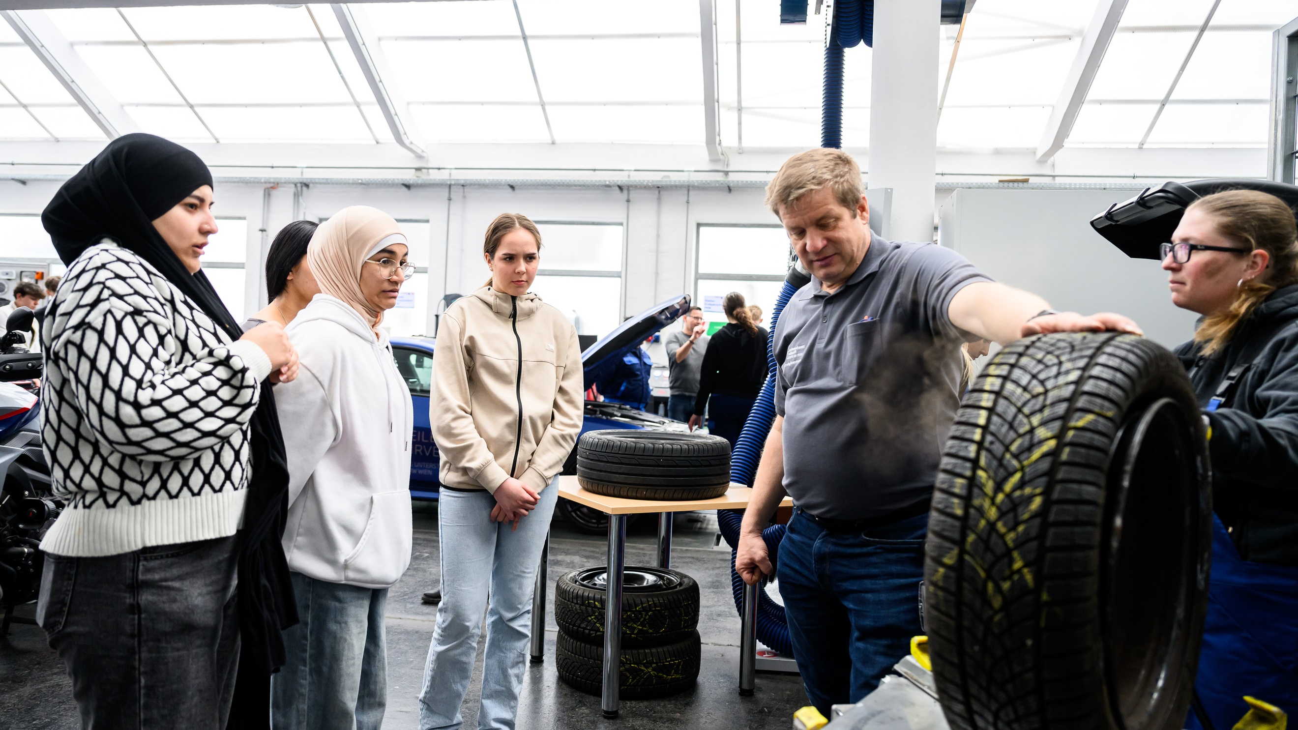 In einer Werkstatt steht ein Autoreifen auf einem Tisch und wird von einer erwachsenen Person mit der Hand festgehalten. Der Person gegenüber sind zwei Teenager und zwei erwachsene Personen