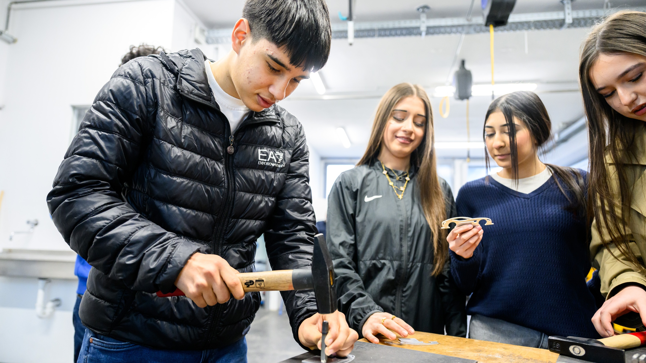 Ein Teenager steht an einem Tisch und schlägt mit einem Hammer einen breiten Nagel in eine MEtallplatte. Daneben stehen drei weitere Teenager