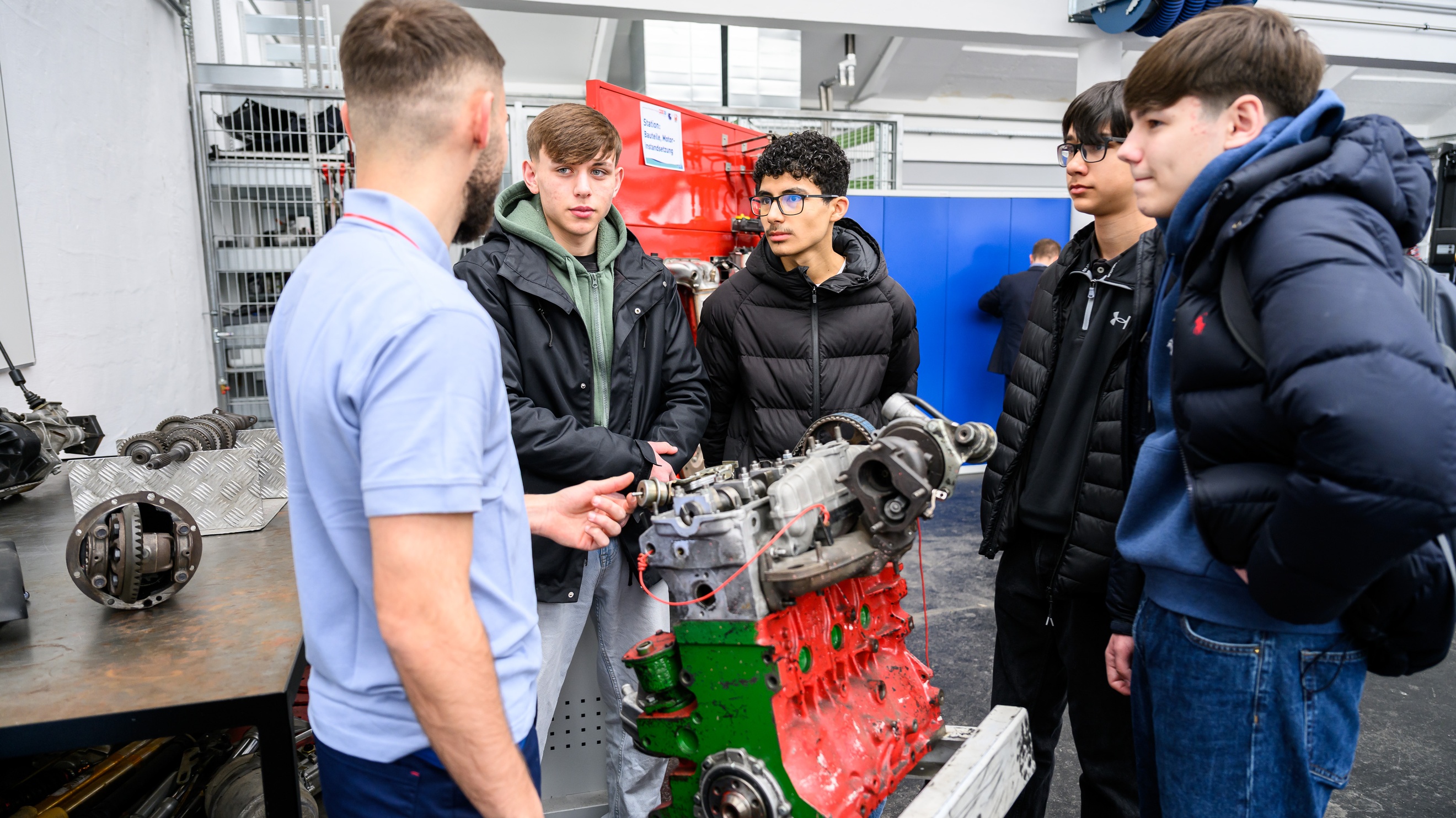 Ein Motor eines Fahrzeugs steht in einer Werkstatt. Um den Motor stehen vier Teenager und eine erwachsene Person. Die Teenager blicken zur erwachsenen Person