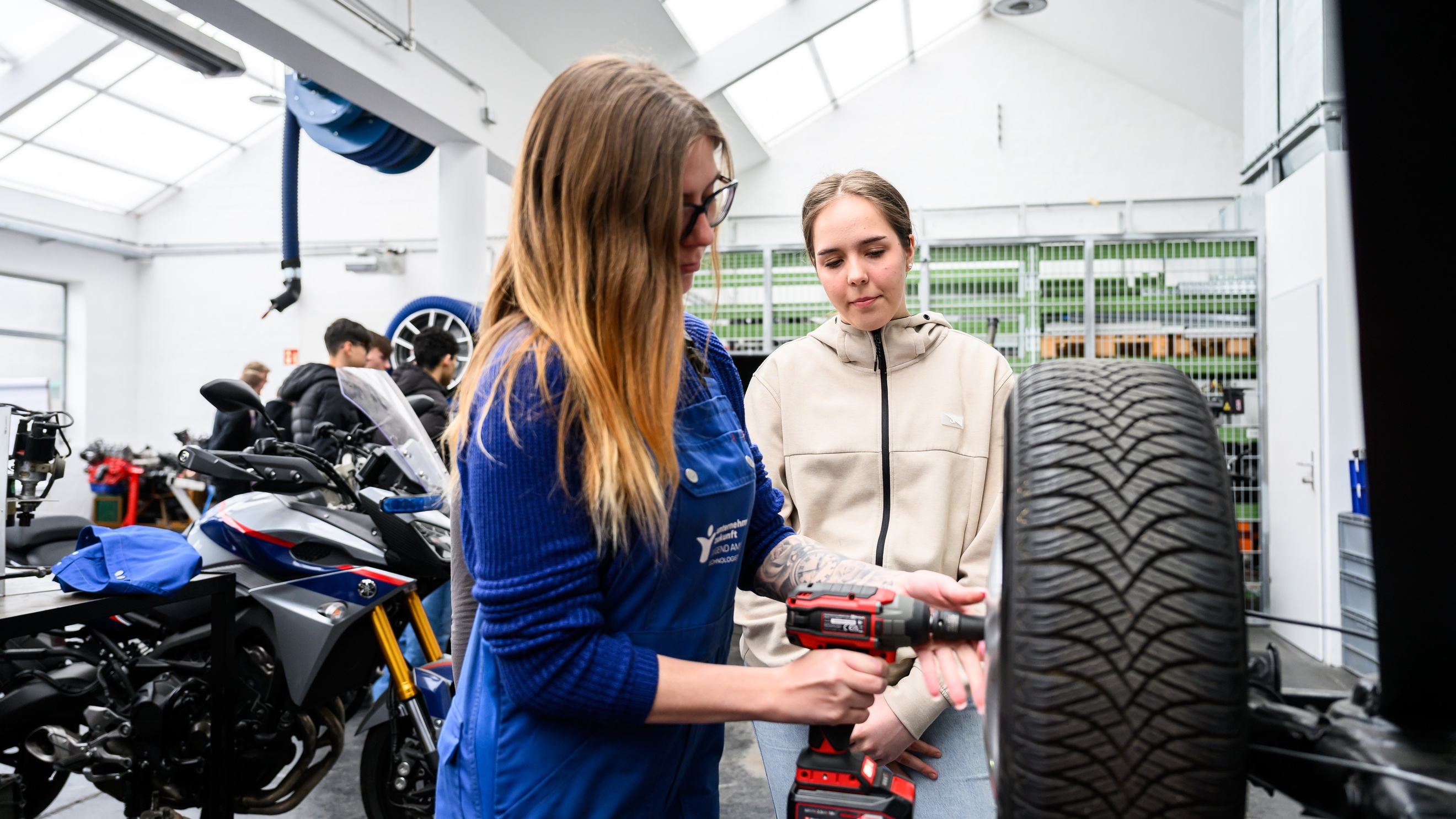  In einer Werkstatt steht ein Autoreifen erhöht. Eine Person fixiert eine Felge in der Mitte des Reifens. Neben der Person steht ein Teenager. Im Hintergrund stehen Motorräder