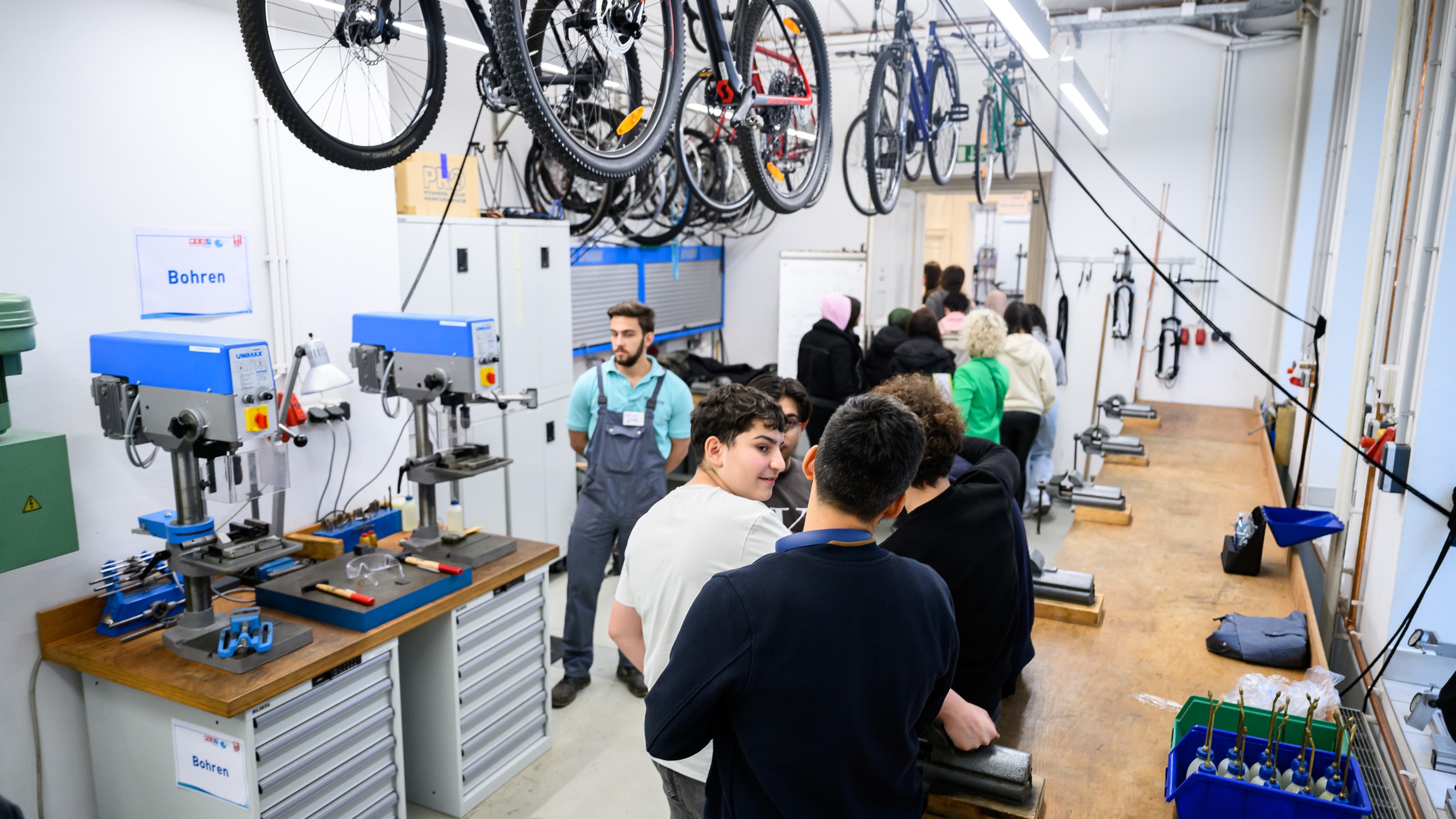 In einem Raum mit Drechslermaschinen stehen viele Jugendliche neben einer Holztischplatte. Neben einer Drechslermaschine steht eine Person in Arbeitskleidung