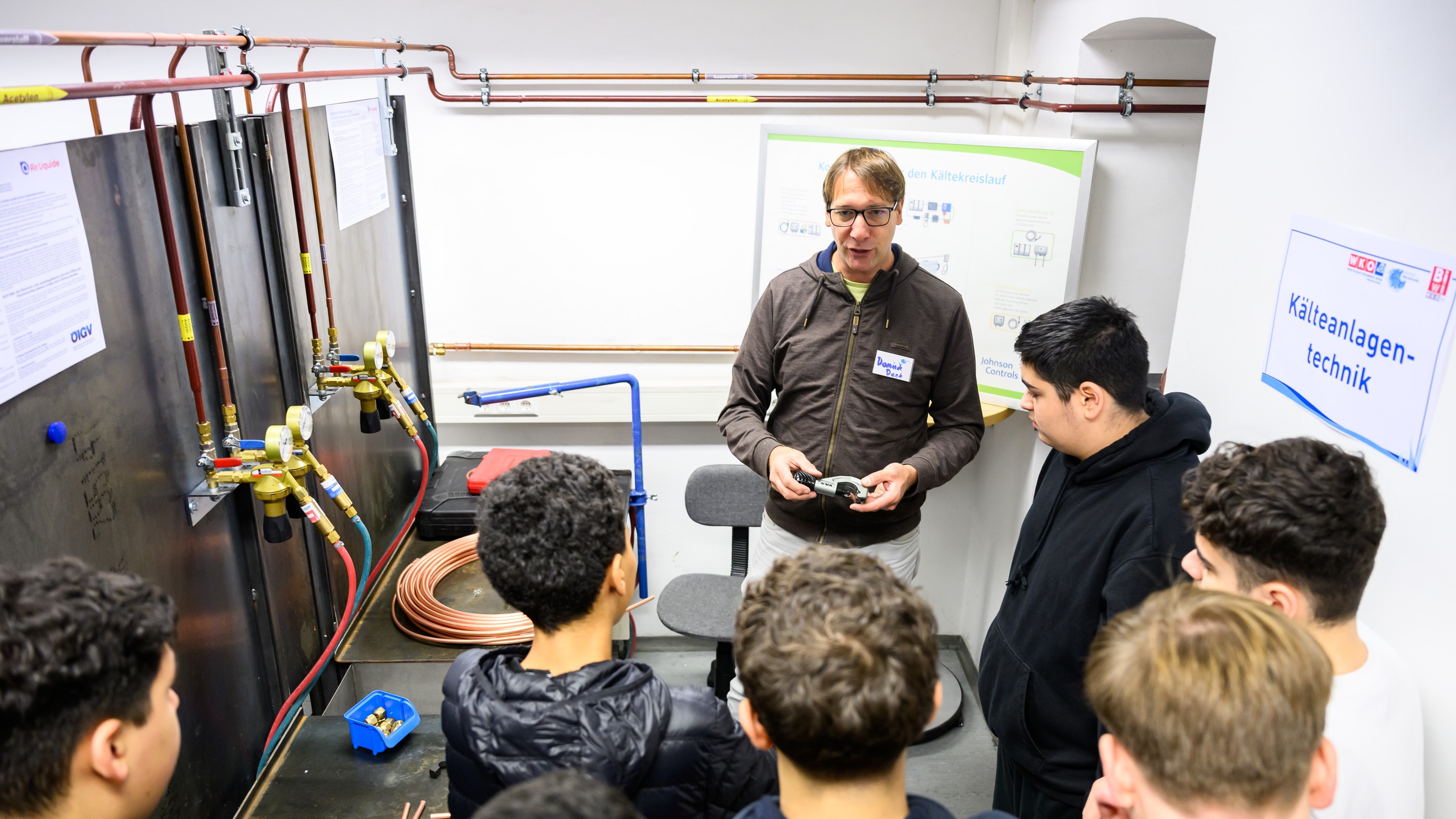 In einem Raum verlaufen an einer Wand verschiedene Rohre. Im Raum steht eine Gruppe Jugendlicher. Ihnen gegenüber ist eine weitere Person, die spricht. An der Wand hängt ein Zettel, auf dem Kälteanlagentechnik steht