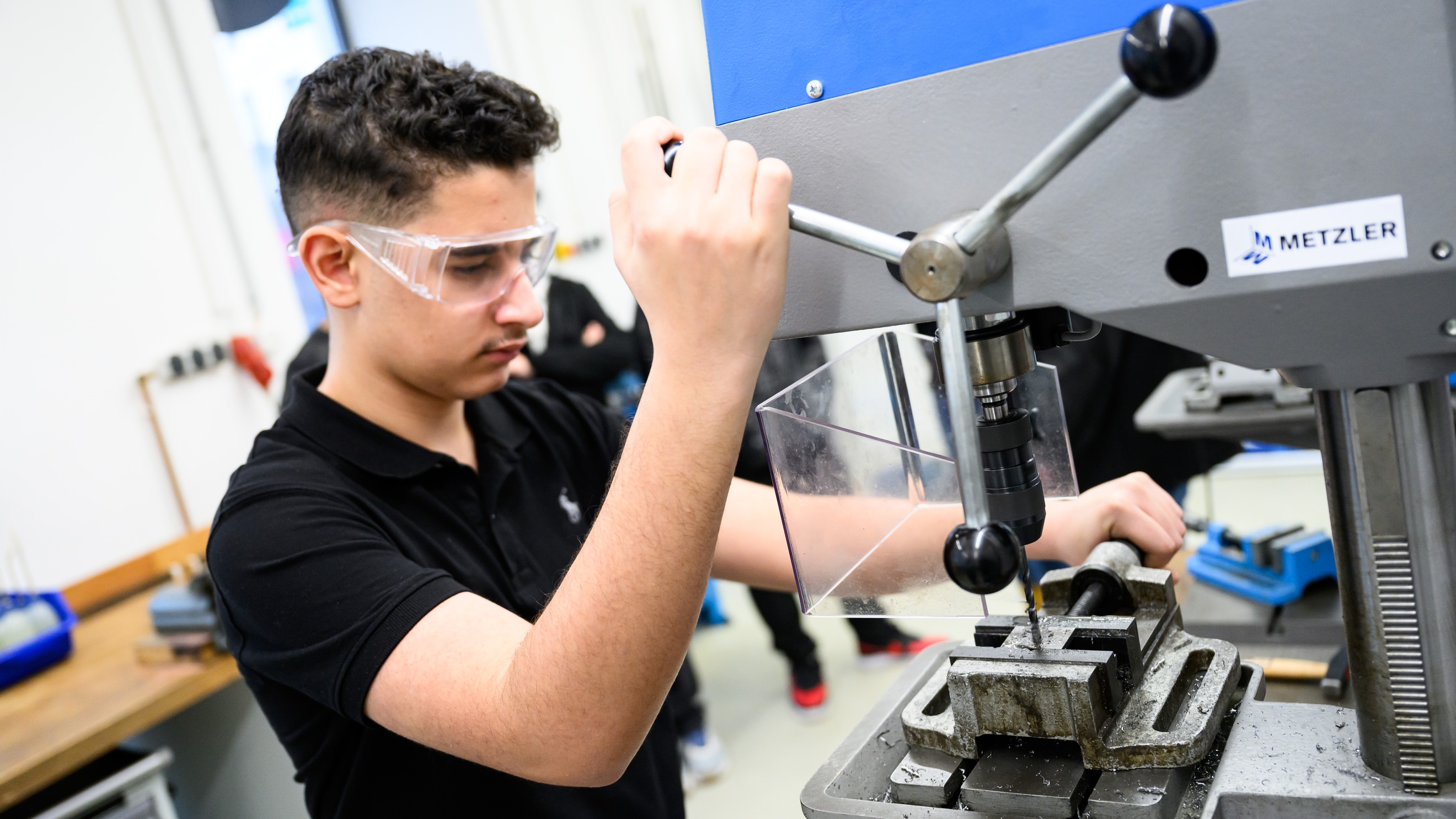  Eine Person mit Schutzbrille steht neben einer Bohrmaschine und betätigt diese