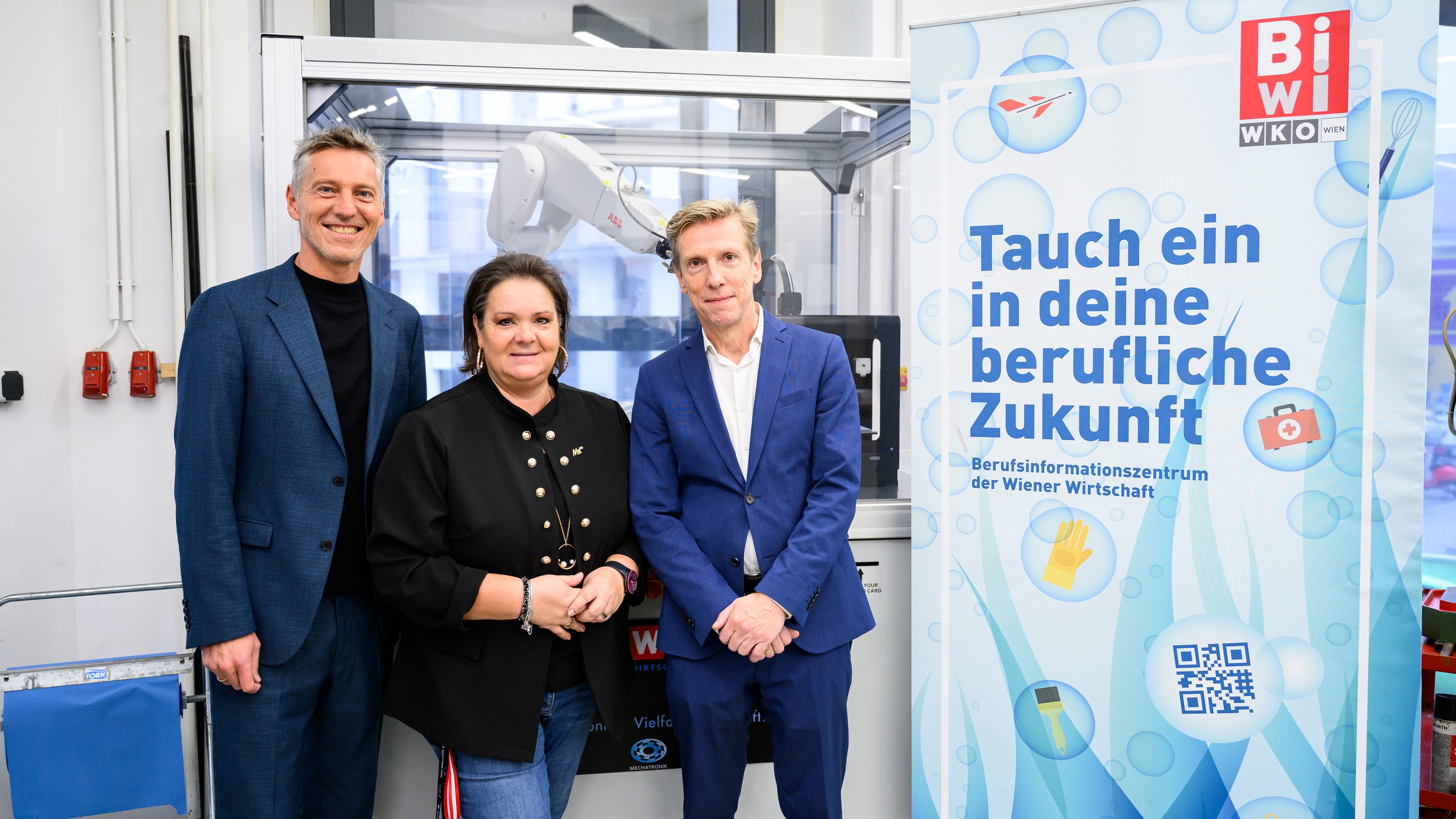 Porträt von drei Personen direkt nebeneinander, die neben einem länglichen Plakat in einem Raum stehen, auf dem Tauch in deine berufliche Zukunft ein steht