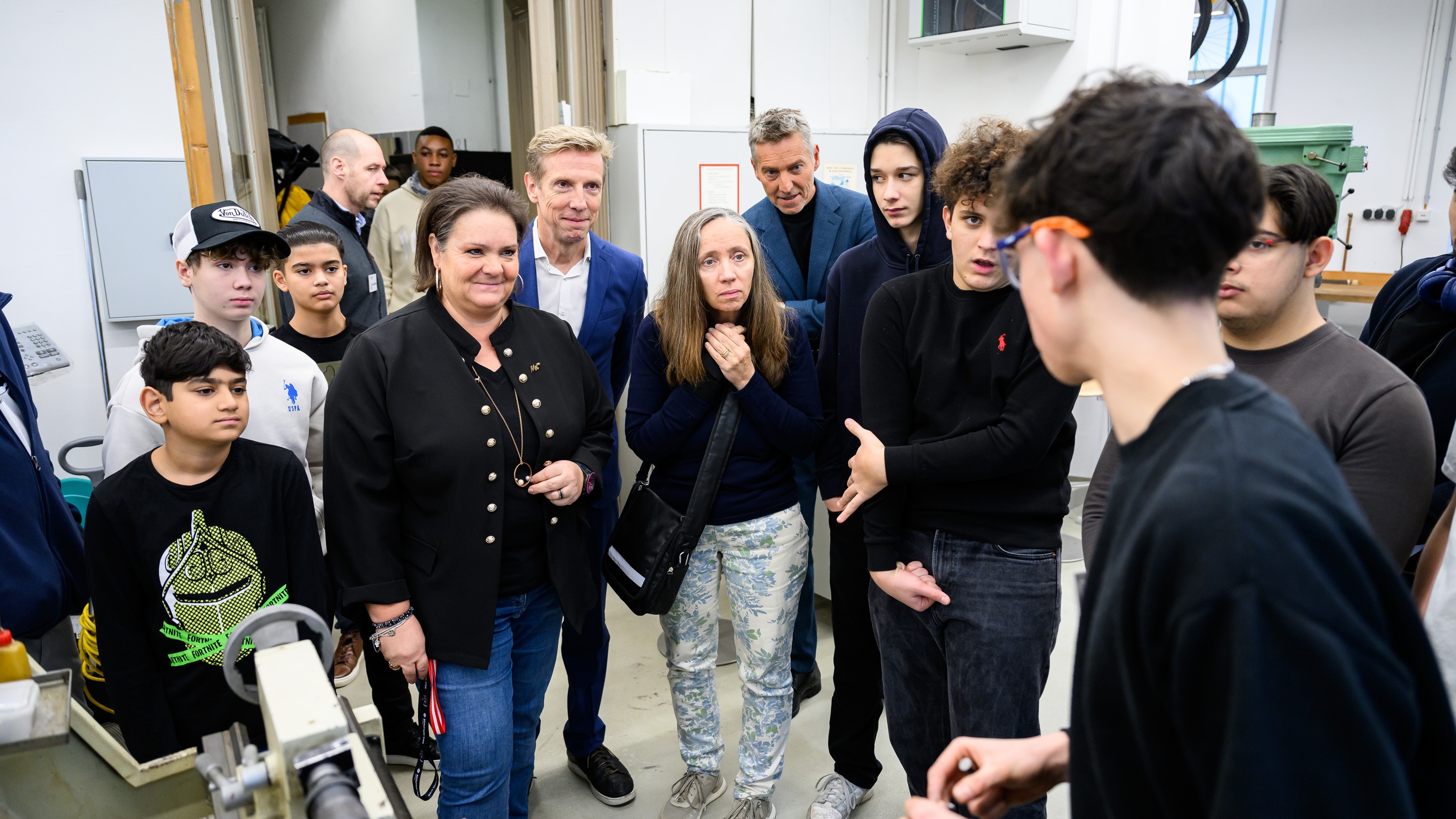 Person mit Schutzbrille steht neben einem Gerät. Ihr gegenüber stehen viele weitere Personen. Die Person mit der Schutzbrille redet zu den anderen Personen