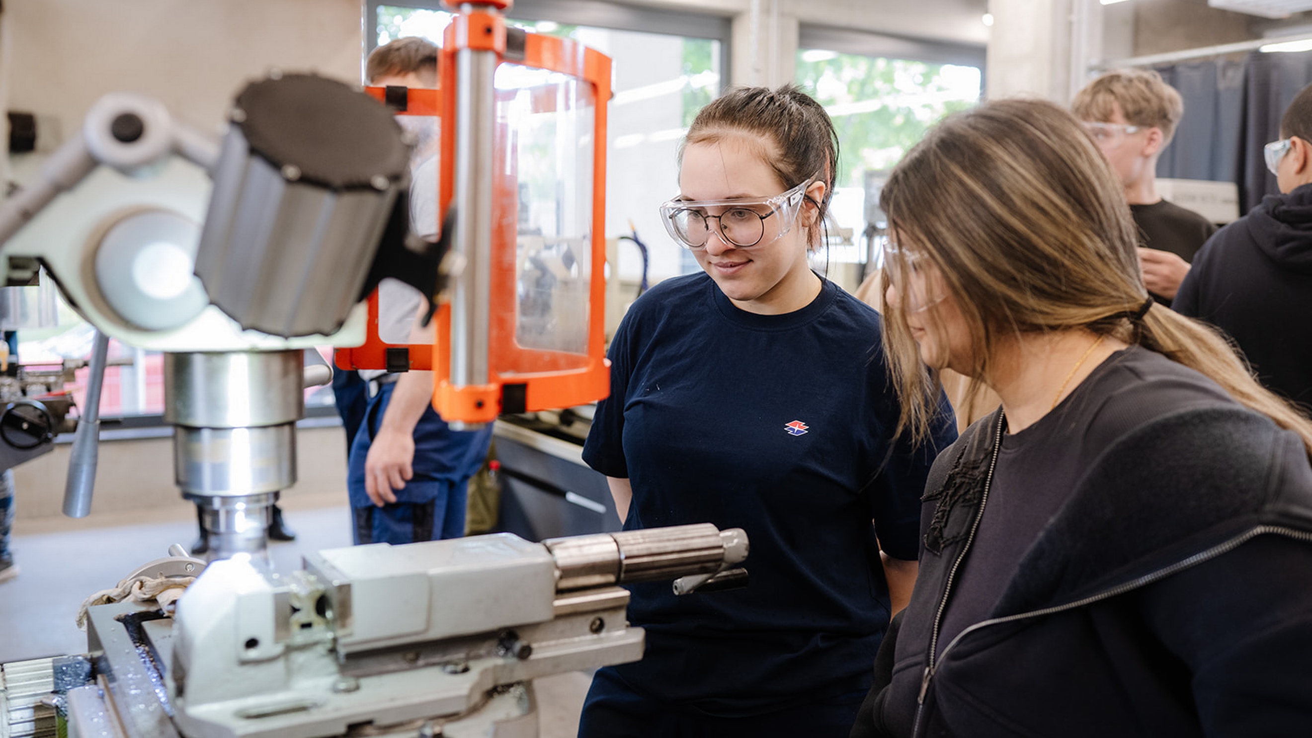 Zwei Personen stehen vor einer Werkzeugmaschine mit transparentem Schutzschild in einer Werkstatt. Beide tragen Schutzbrillen und blicken auf den Arbeitsbereich der Maschine. Im Hintergrund befinden sich weitere Personen