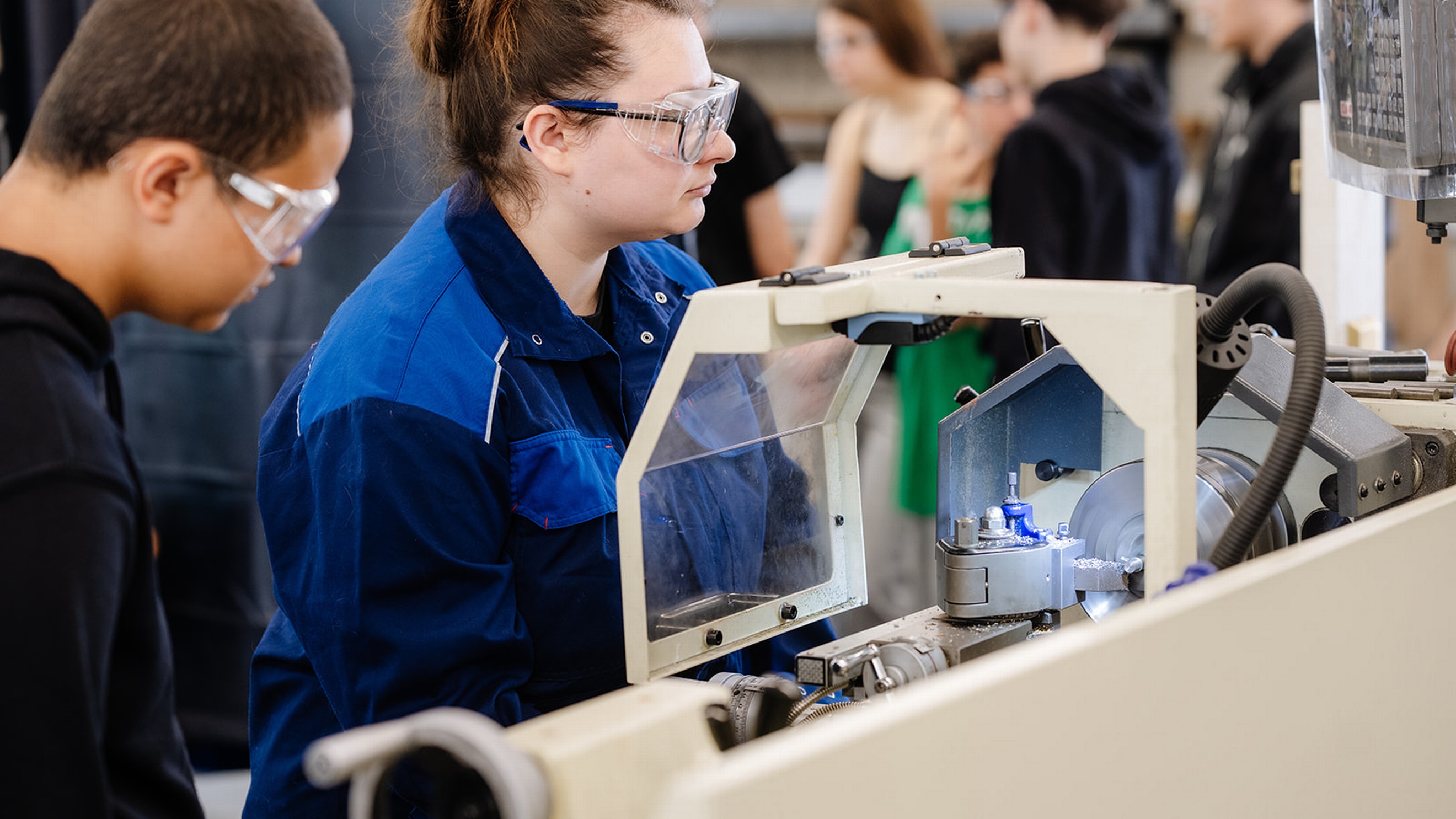 Seitliche Aufnahme von zwei Personen, die nebeneinander an einer Maschine mit Schutzabdeckung stehen. In der Maschine ist ein Metallstück eingespannt