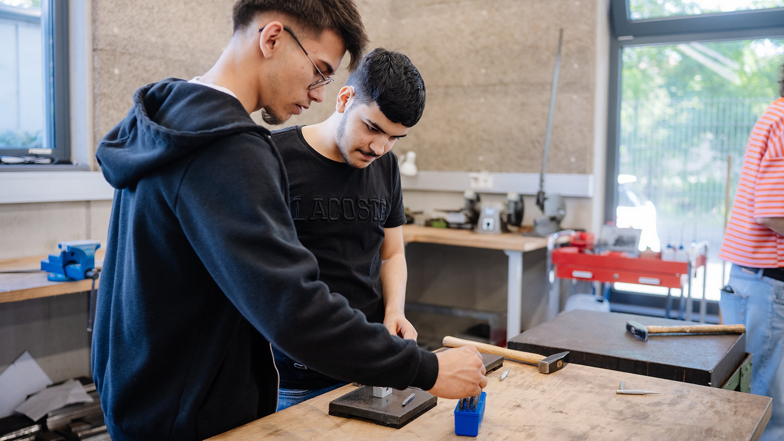 Zwei Personen stehen an einer Werkbank in einer Werkstatt. Auf der Arbeitsfläche liegen ein Hammer, Metallstifte und weitere Werkzeuge