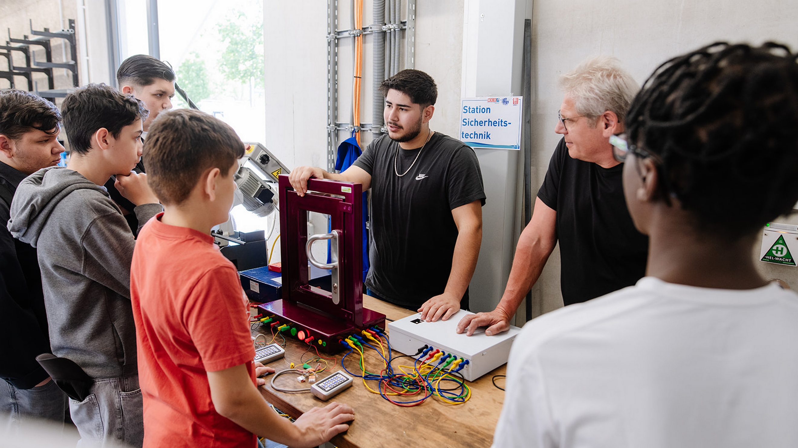 Mehrere Personen stehen um eine Werkbank mit elektronischen Komponenten und Kabelverbindungen. Zwei Personen stehen an einer technischen Anwendung mit einem Türrahmen-Modell und angeschlossenen Geräten. Im Hintergrund hängt ein Schild mit der Aufschrift „Station Sicherheitstechnik“