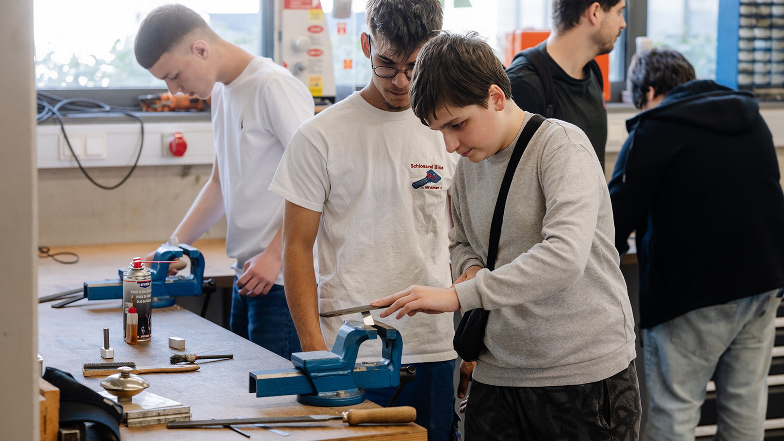 Mehrere Personen arbeiten in einer Werkstatt an Werkbänken mit Schraubstöcken und Werkzeugen. Im Vordergrund bearbeitet eine Person ein eingespanntes Metallstück mit einer Feile. Weitere Personen stehen an benachbarten Arbeitsplätzen. Auf der Werkbank liegen Werkzeuge und Materialien