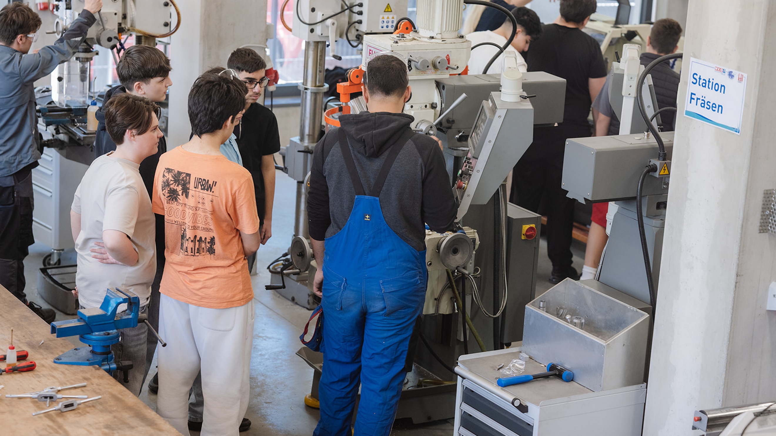 Mehrere Personen stehen in einer Werkstatt um eine Fräsmaschine und beobachten bzw. bedienen die Anlage. Eine Person in blauer Arbeitskleidung arbeitet direkt an der Maschine. Im Vordergrund befinden sich Werkzeuge und ein Schraubstock auf einer Werkbank