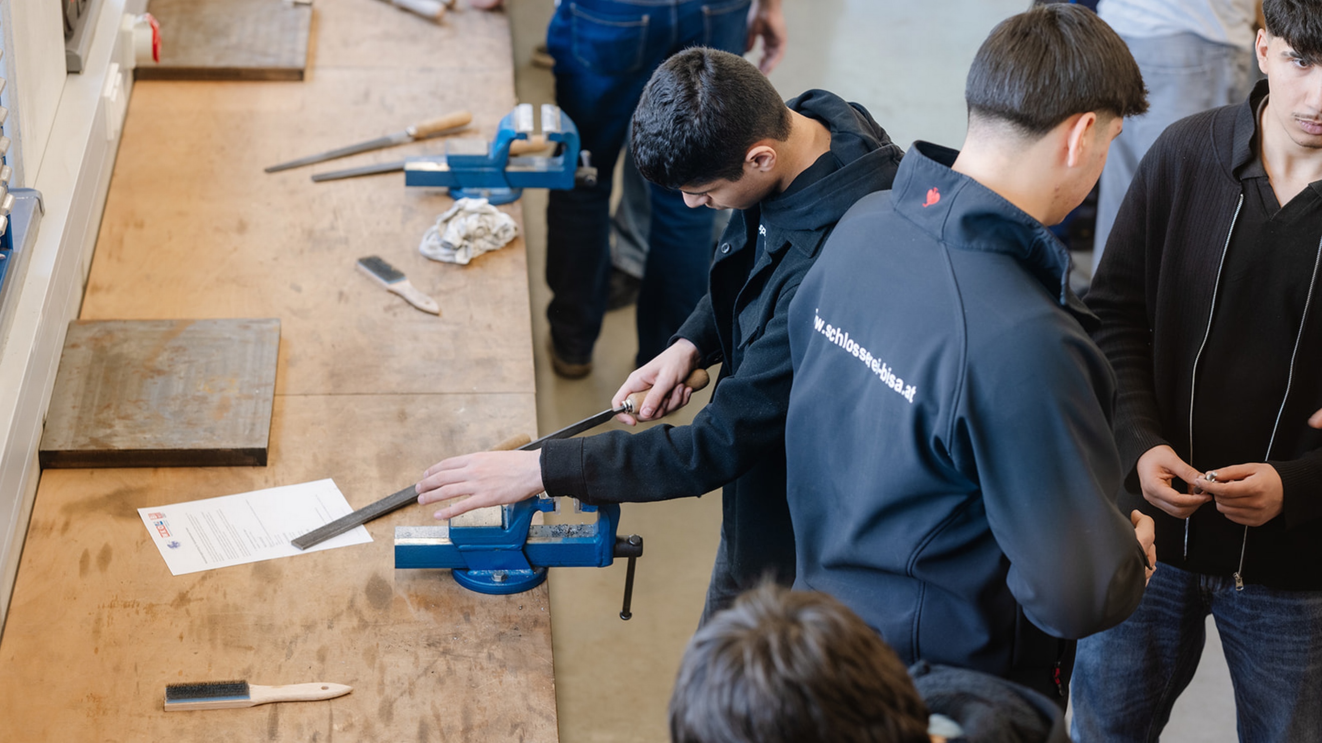Mehrere Personen stehen an einer langen Werkbank in einer Werkstatt. Eine Person bearbeitet ein eingespanntes Metallteil mit einer Feile in einem blauen Schraubstock. Auf der Werkbank liegen weitere Feilen, Werkzeuge und Metallteile