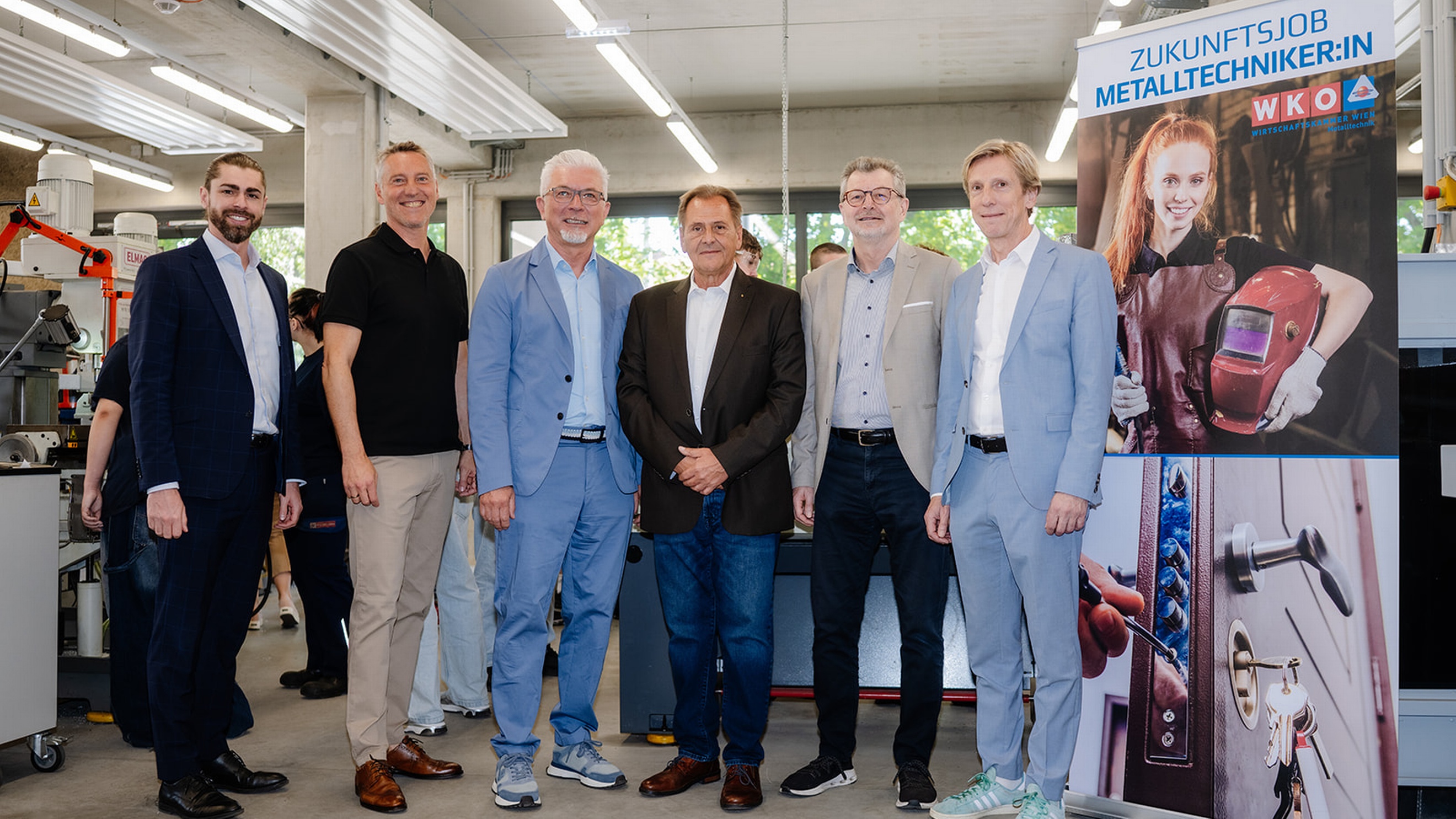 Sechs Personen stehen in einer Werkstatt mit Maschinen und Arbeitsplätzen. Rechts befindet sich ein Roll-up mit der Aufschrift „Zukunftsjob Metalltechniker:in“. Das Roll-up zeigt eine Person mit Schweißhelm sowie ein Detailbild eines Türschlosses mit Schlüssel