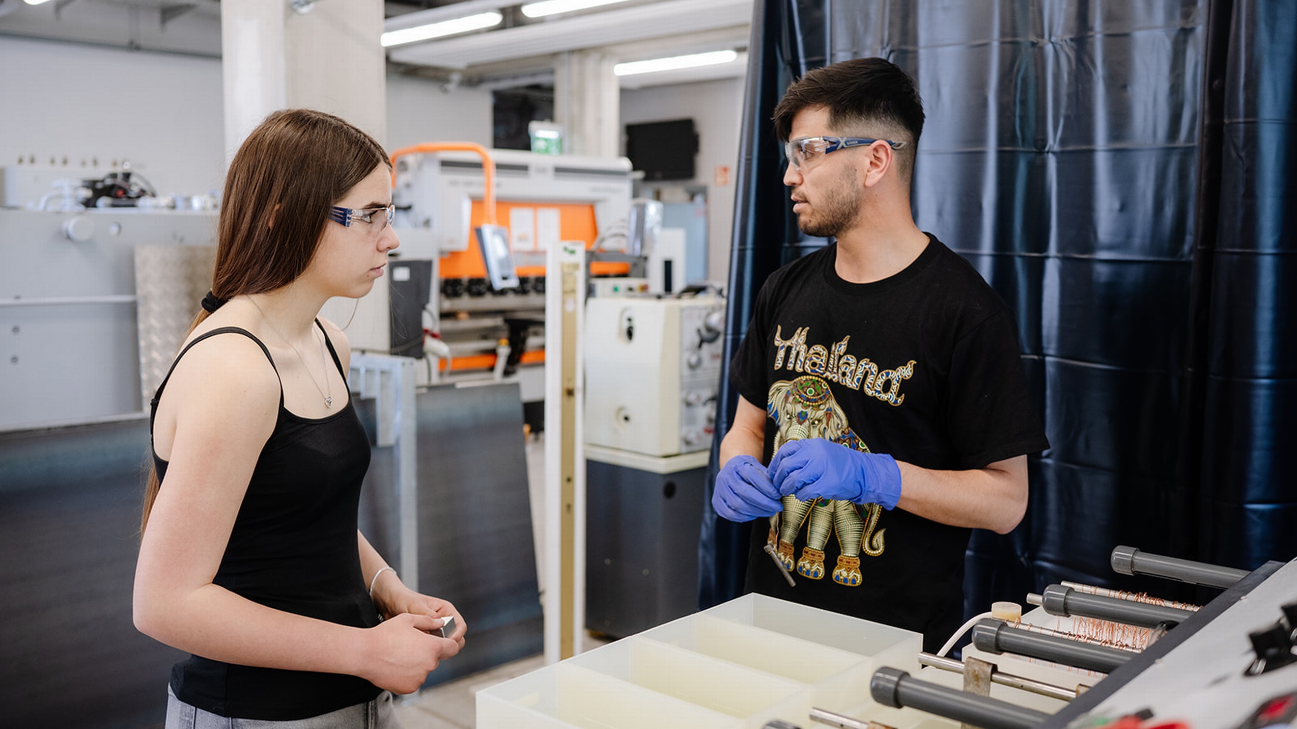 Zwei Personen stehen in einer Werkstatt neben einer technischen Anlage. Beide tragen Schutzbrillen, eine Person zusätzlich blaue Schutzhandschuhe. Im Vordergrund befindet sich ein Maschinenteil mit Metallrollen und Kupferdraht