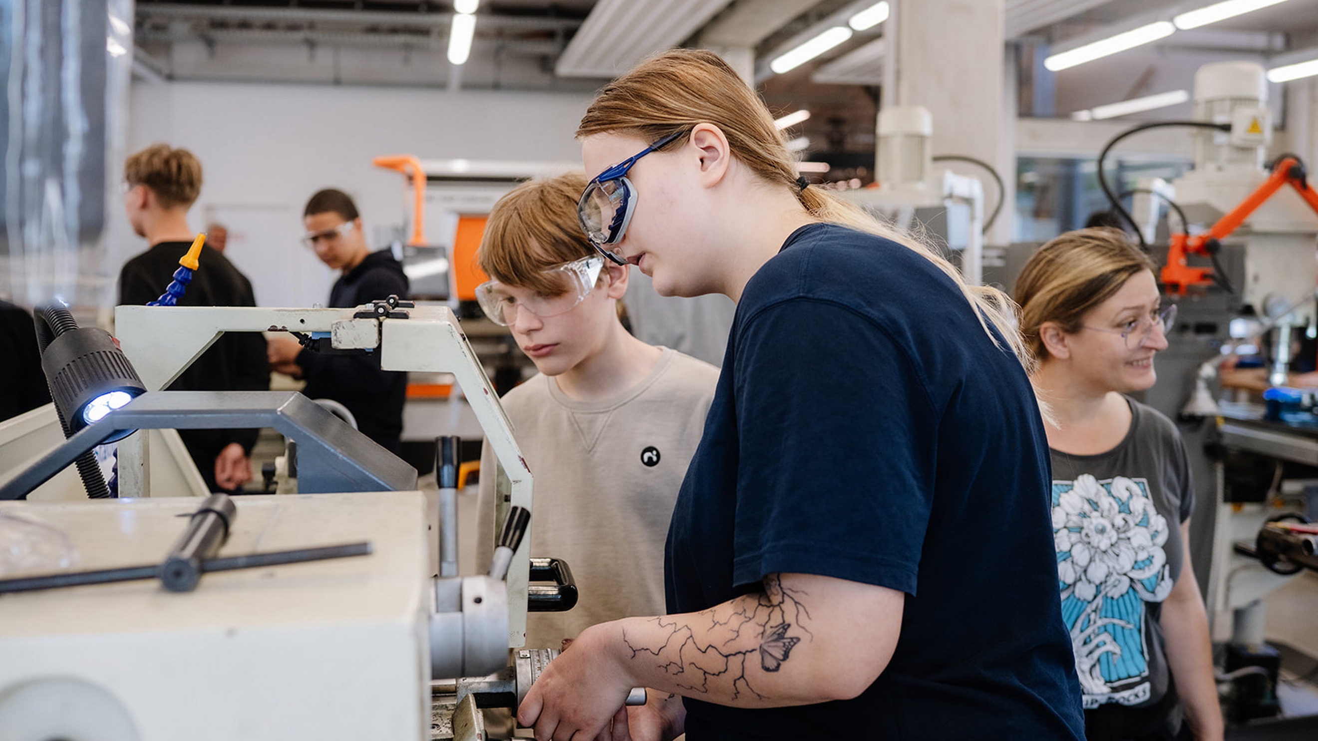 Mehrere Personen arbeiten in einer Werkstatt an einer Maschine. Zwei Personen im Vordergrund tragen Schutzbrillen und bedienen gemeinsam eine Anlage