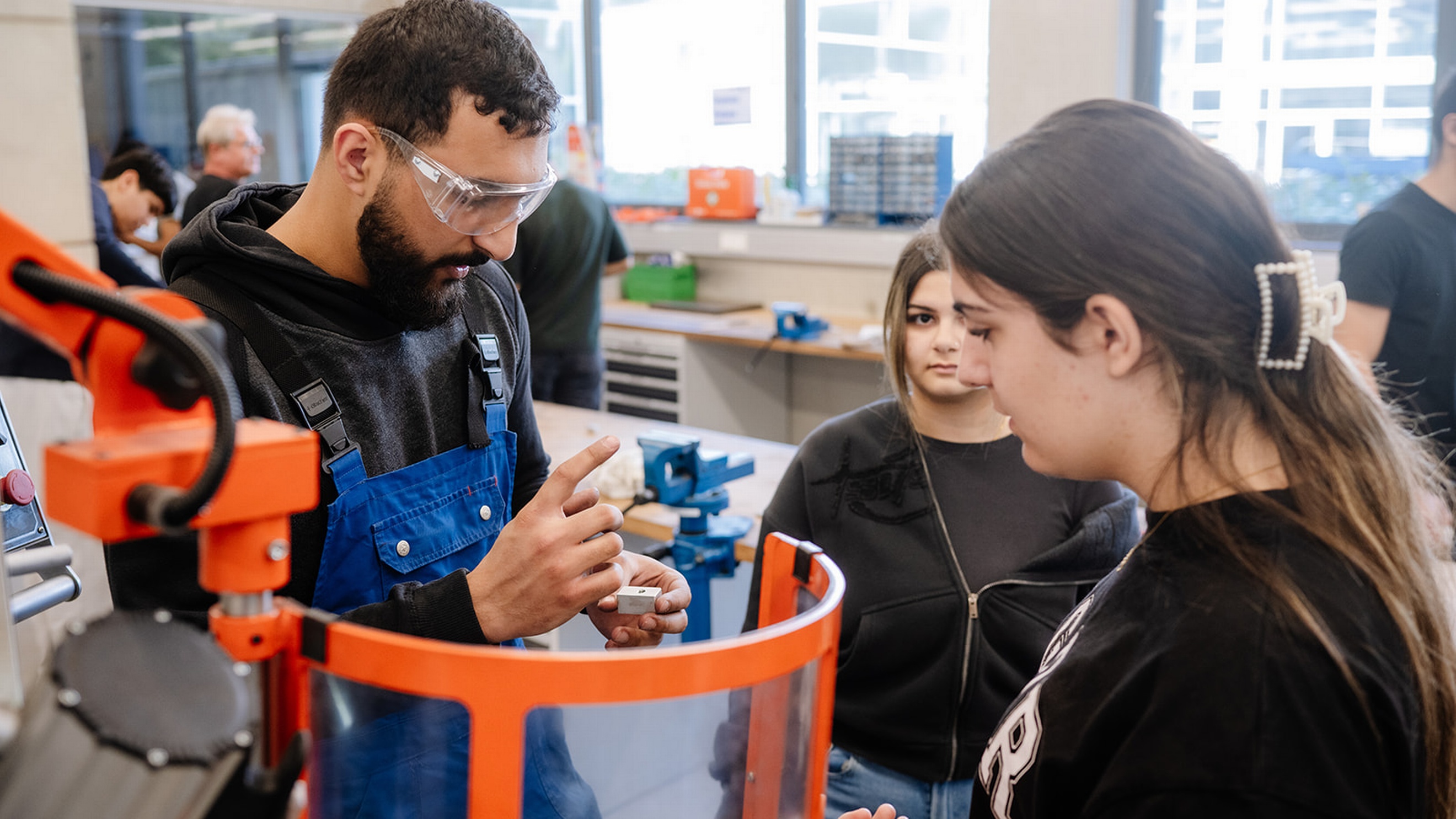 Eine Person mit Schutzbrille und Arbeitskleidung hält ein kleines Werkstück neben einer Maschine mit transparentem Schutzschild. Ihr gegenüber stehen zwei weitere Personen. Eine der Personen blickt auf das Werkstück. Im Hintergrund befinden sich Werkbänke, Schraubstöcke und weitere Personen