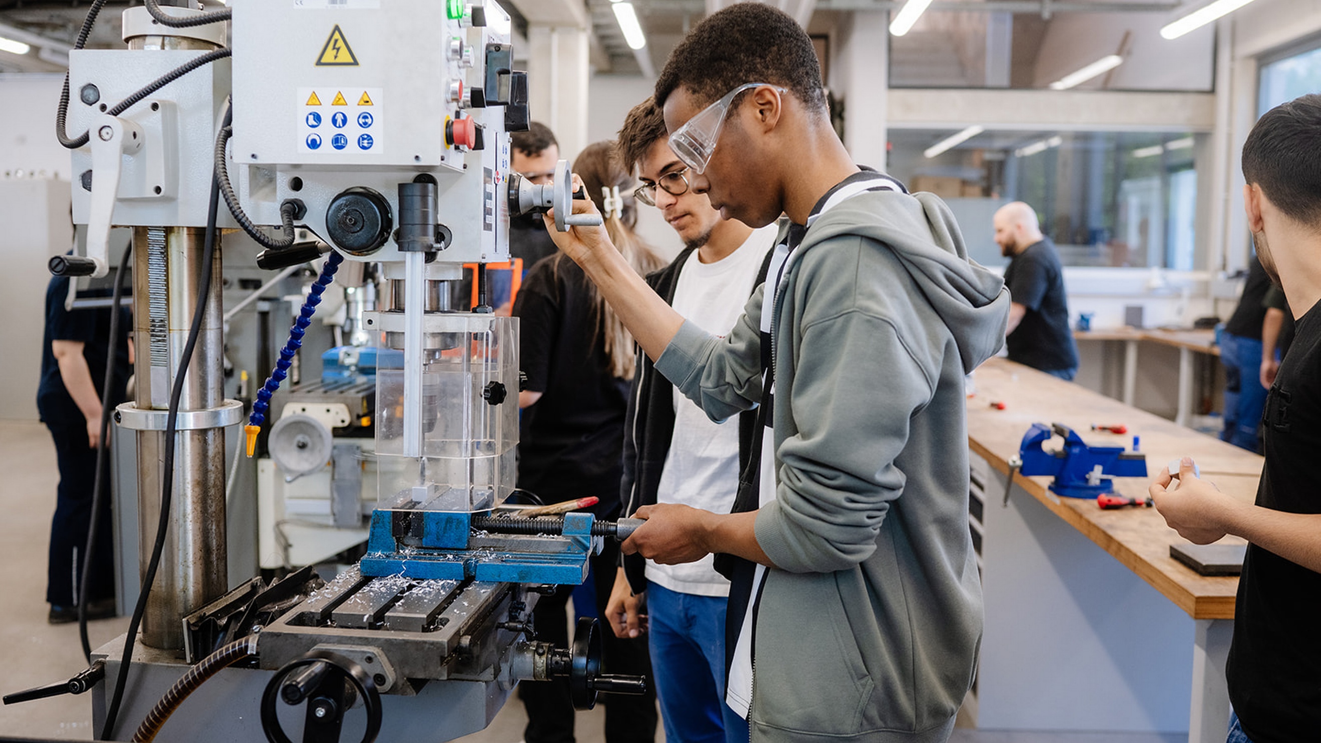 Mehrere Personen stehen an einer Fräsmaschine in einer Werkstatt. Zwei Personen bedienen die Maschine und halten Werkstück sowie Bedienelemente. Beide tragen Schutzbrillen. Auf dem Arbeitstisch der Maschine liegen Metallspäne