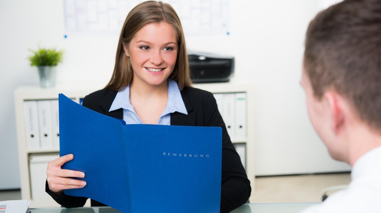 Zwei Personen sitzen sich an einem Tisch gegenüber. Die zur Kamera gewandte Person hält eine blaue Mappe, auf der Bewerbung steht. Vor ihr sind ein Notizblock und ein Stift